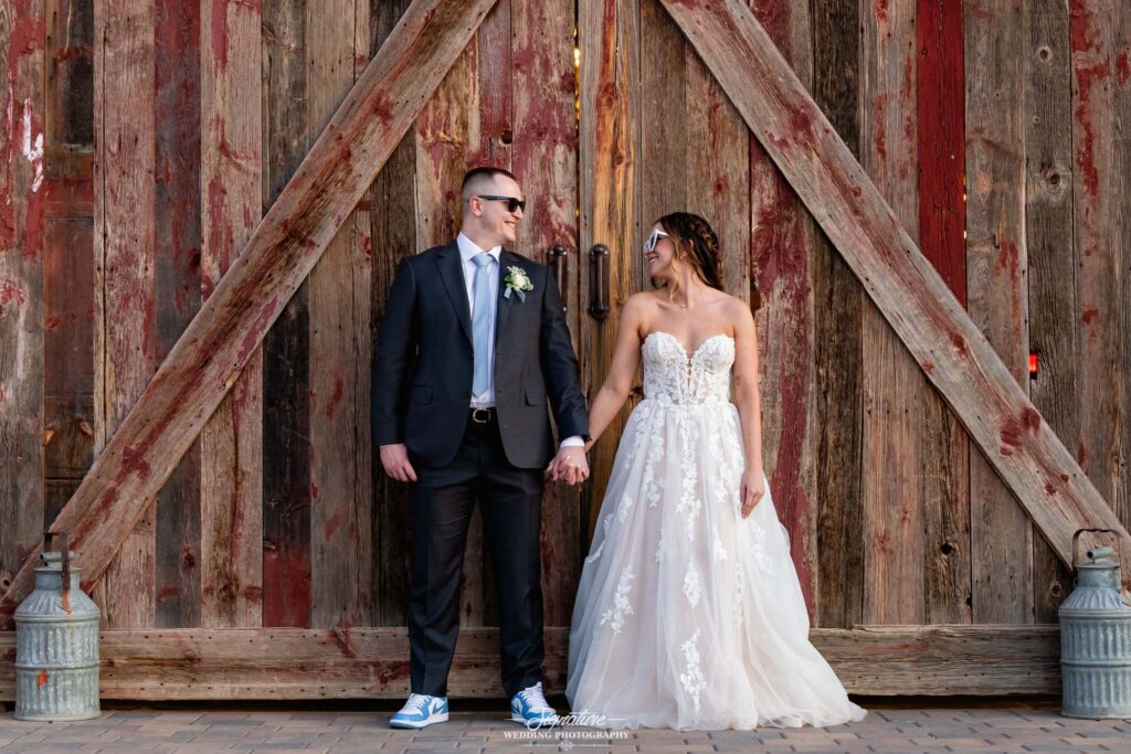 Image by Signature Wedding Photography A couple in wedding attire stands holding hands in front of a large rustic wooden door. The groom wears a suit with sneakers, and the bride wears a strapless lace gown. They face each other and smile.