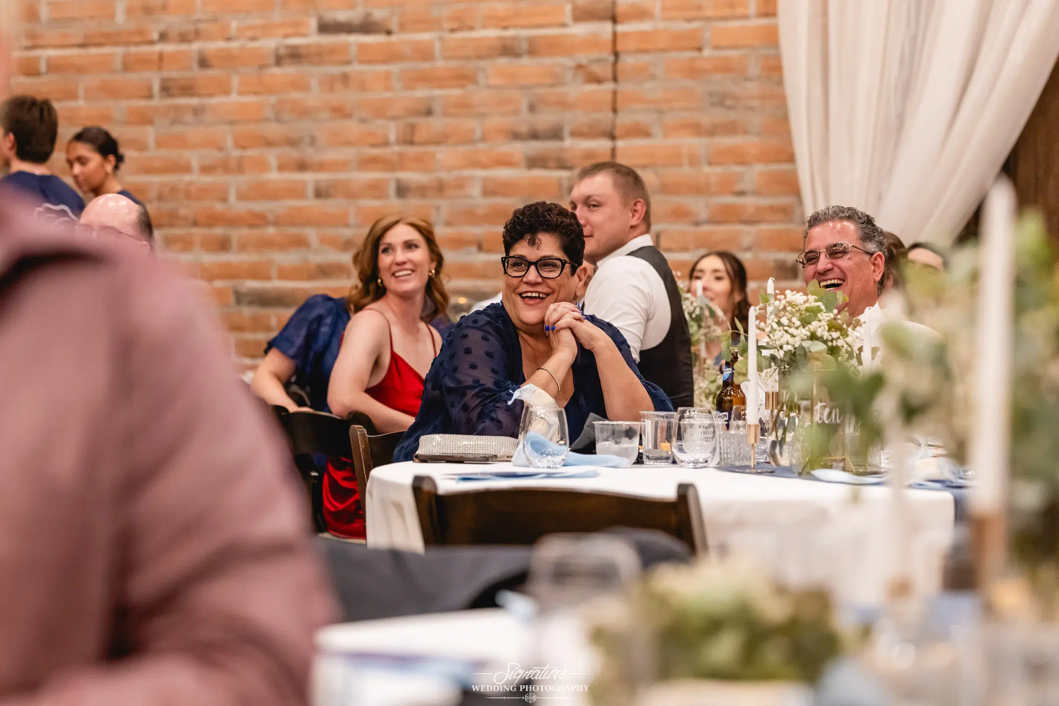 Image by Signature Wedding Photography Guests sit at decorated tables, smiling and watching something off-camera at an indoor event with a brick wall backdrop. Some people are dressed formally, and the atmosphere appears joyful and celebratory.