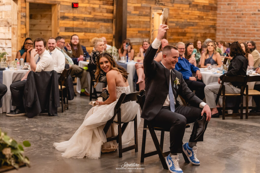 A bride and groom, sitting back-to-back, each hold up a shoe during their wedding reception game. Guests watch and smile in a rustic venue with wooden walls and warm lighting.