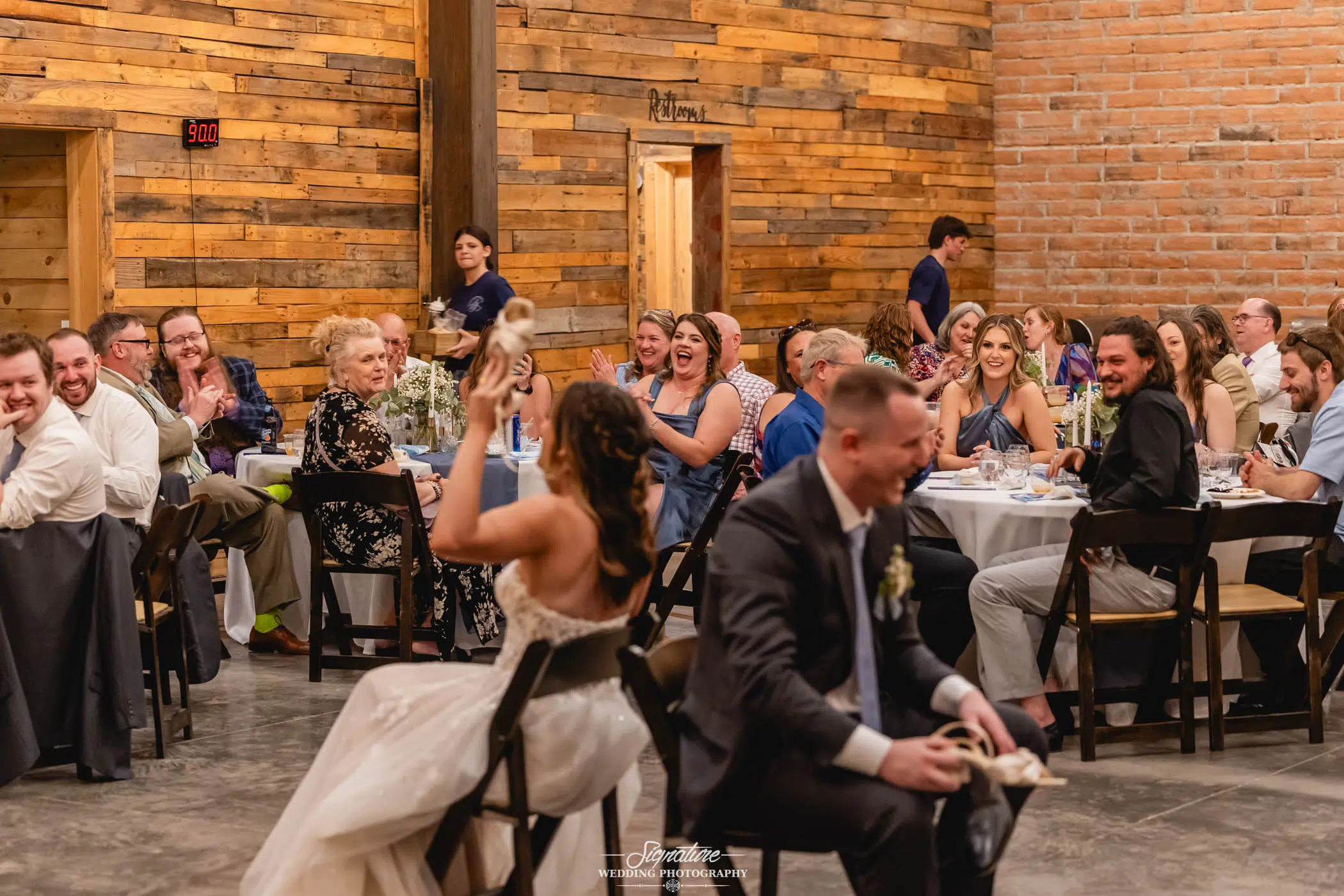 Image by Signature Wedding Photography A bride and groom play the shoe game at their wedding reception, sitting back-to-back and laughing as guests seated at round tables watch and smile in a rustic wooden hall.