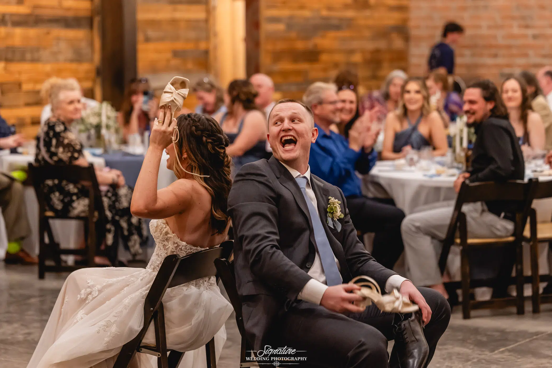 Image by Signature Wedding Photography A bride and groom sit back-to-back holding each other's shoes, laughing, while wedding guests seated at tables in the background watch and smile during a lively reception game.