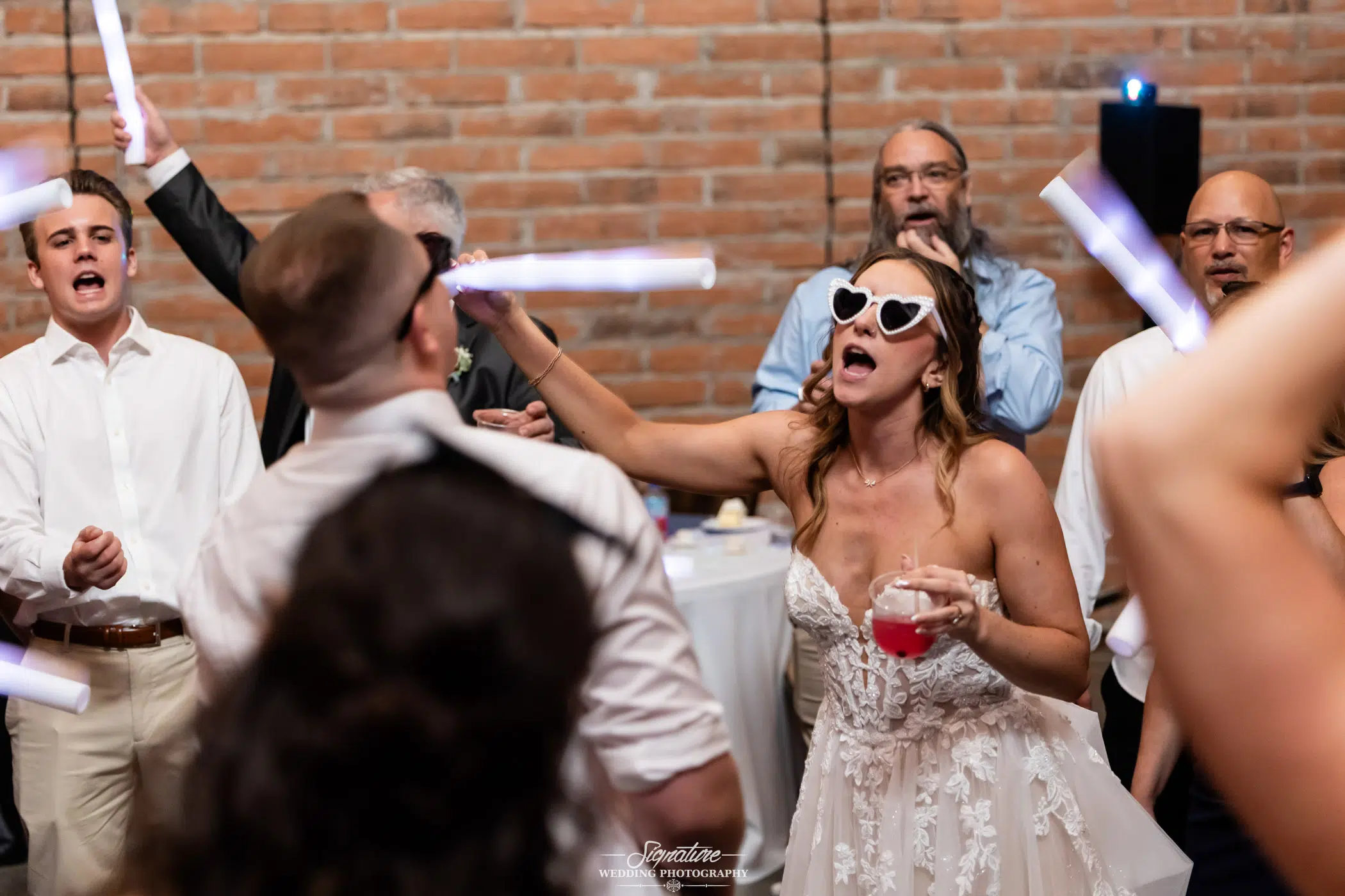 Image by Signature Wedding Photography A bride in a strapless dress and heart-shaped sunglasses dances energetically with guests at a lively wedding reception, holding a drink and a light stick. People around her are singing and celebrating.