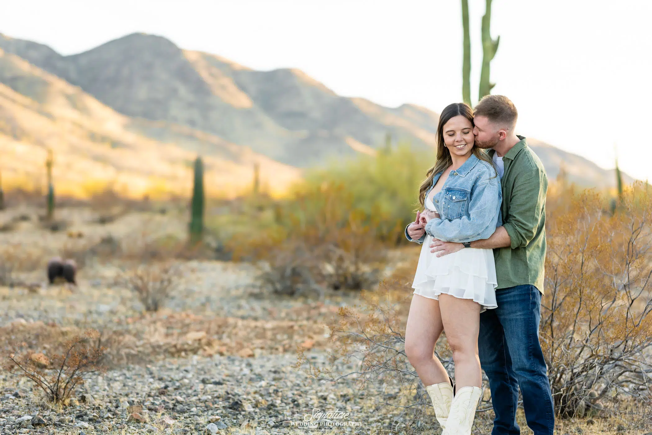 Image by Signature Wedding Photography A couple embraces in a desert landscape with mountains and cacti in the background. The woman wears a denim jacket, white dress, and cowboy boots; the man stands behind her, wearing a green shirt and jeans.