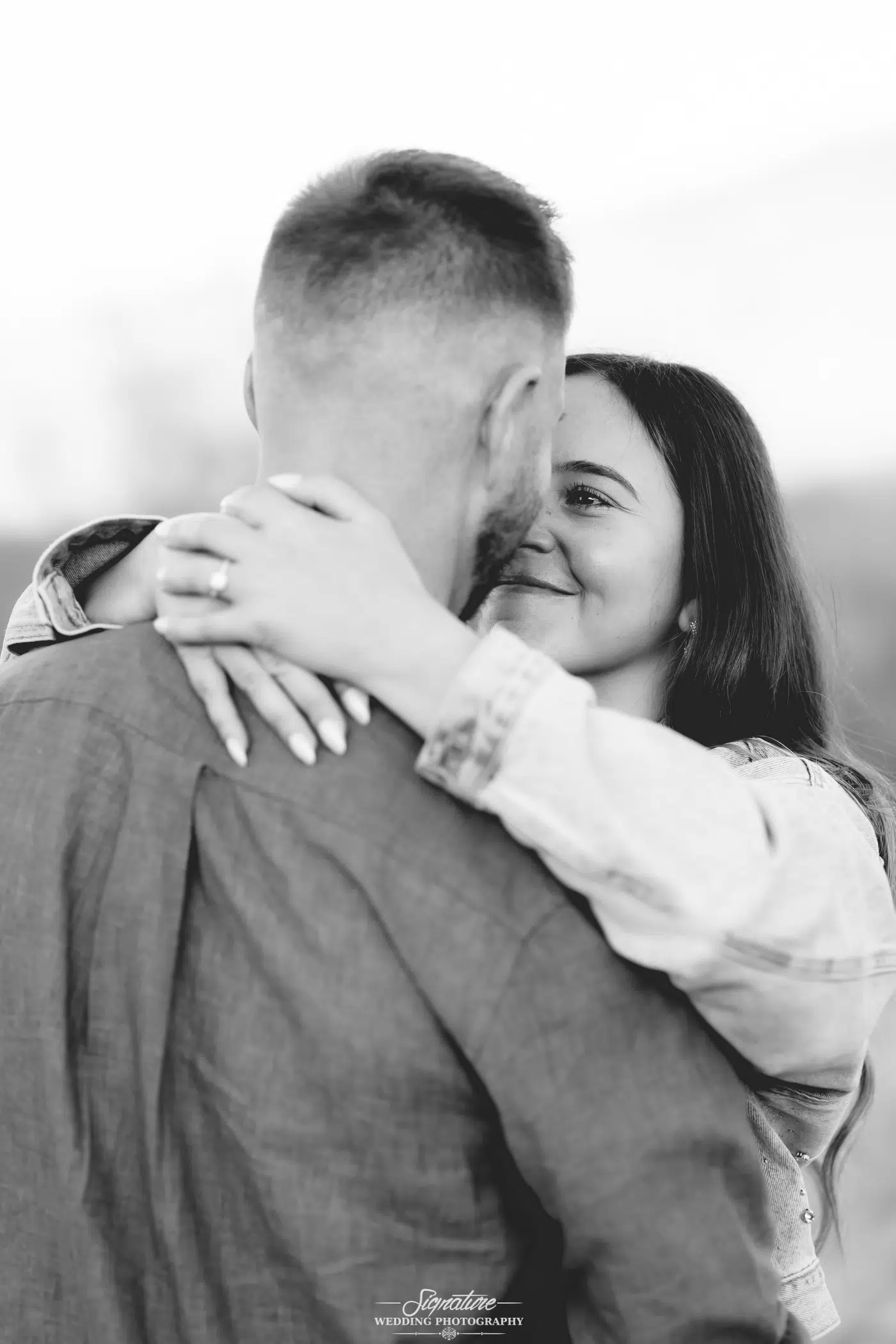 Image by Signature Wedding Photography A woman smiles while embracing a man, her arms around his neck, displaying an engagement ring on her finger. They gaze at each other closely in a loving moment. The photo is in black and white.