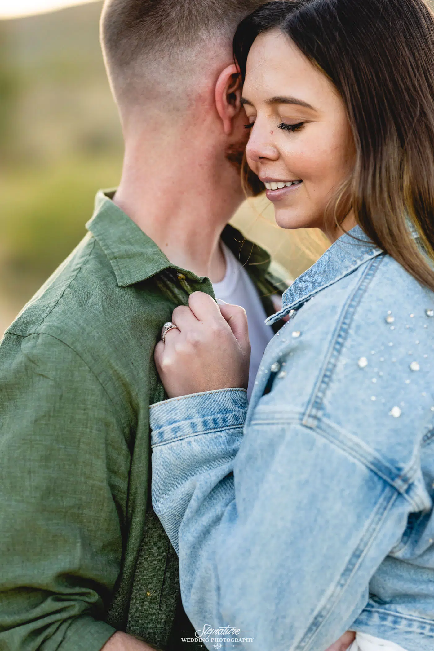 Image by Signature Wedding Photography A woman smiles with her eyes closed, embracing a man closely. She wears a blue denim jacket with pearl details and shows an engagement ring on her hand resting on his chest. The background is softly blurred outdoors.