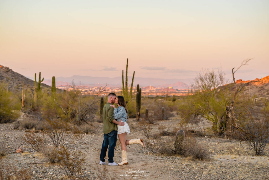 A couple embraces and shares a kiss in a desert landscape at sunset, surrounded by cacti and dry shrubs, with a distant city visible beneath a soft, pastel sky.
