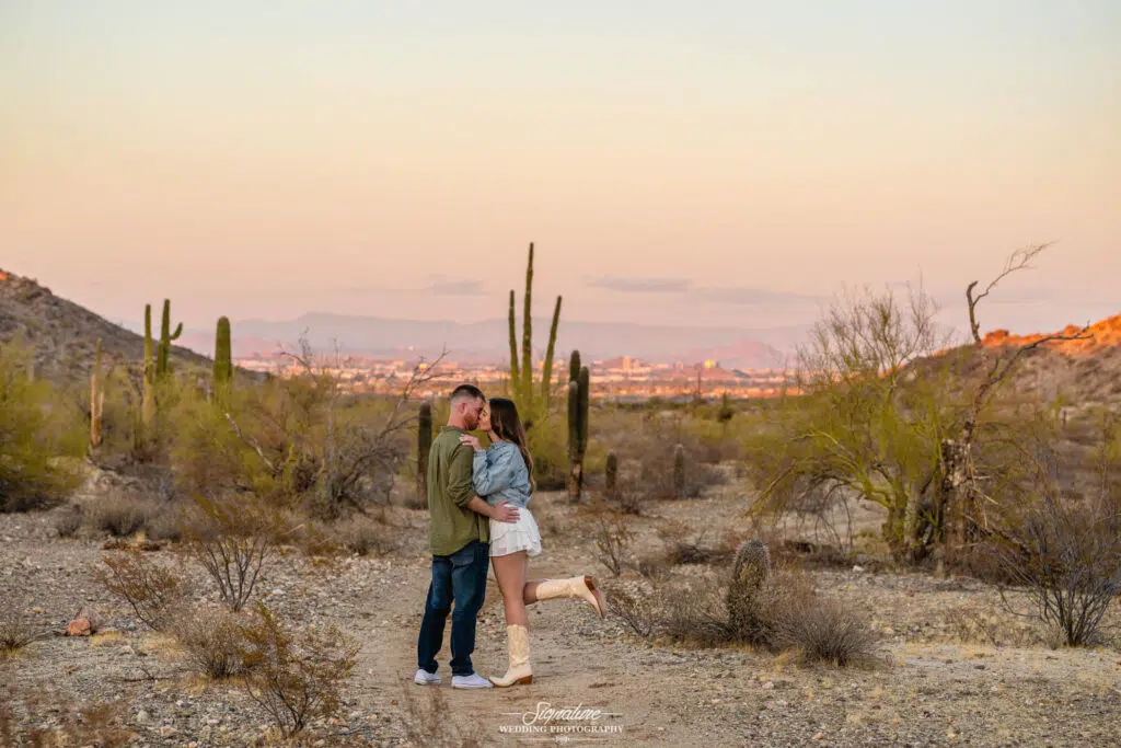 A couple embraces and shares a kiss in a desert landscape at sunset, surrounded by cacti and dry shrubs, with a distant city visible beneath a soft, pastel sky.