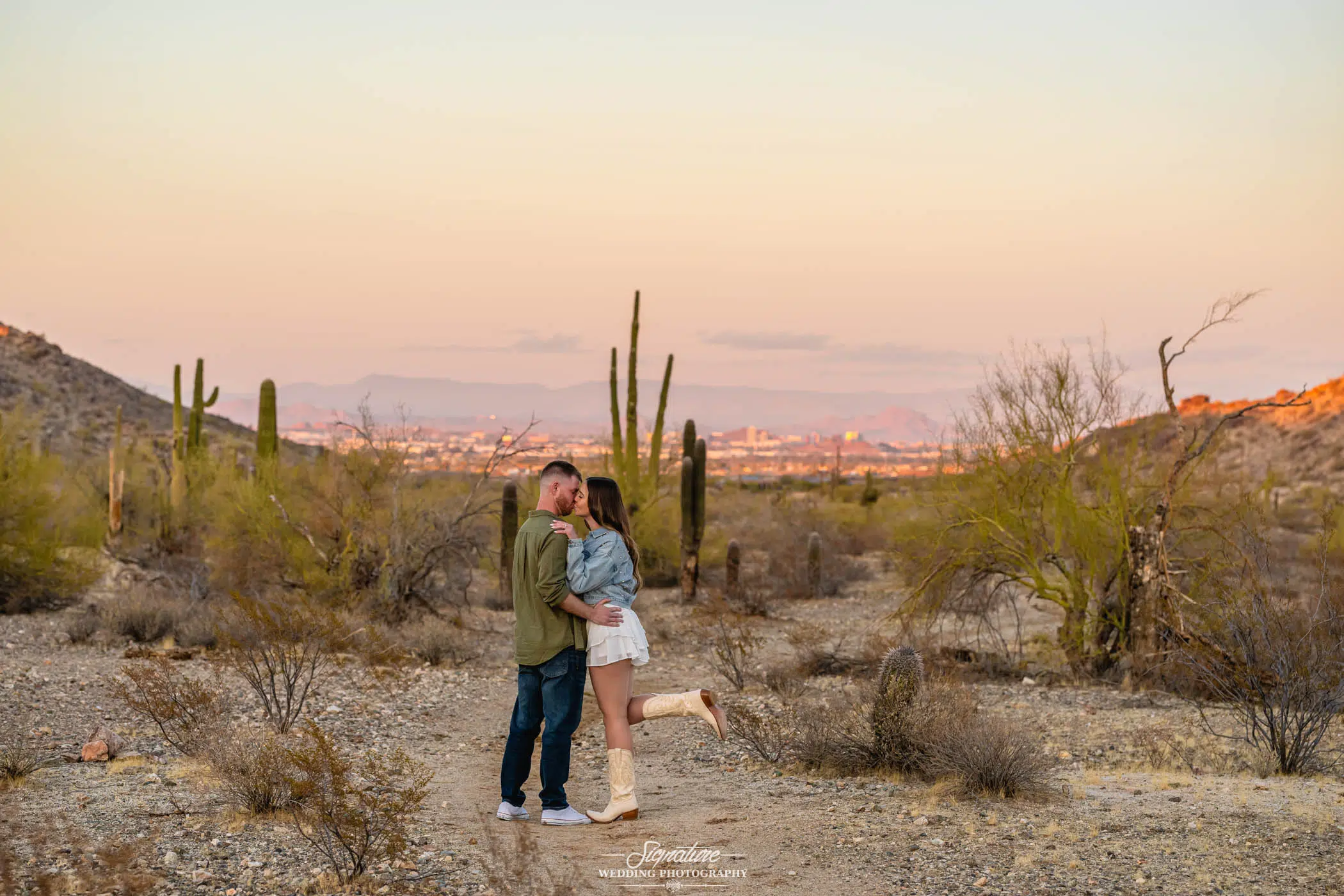 Image by Signature Wedding Photography A couple embraces and shares a kiss in a desert landscape at sunset, surrounded by cacti and dry shrubs, with a distant city visible beneath a soft, pastel sky.