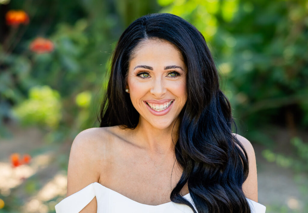 Image by Signature Wedding Photography A smiling bride with long dark hair wears an off-the-shoulder white wedding dress and holds a bouquet of white flowers, standing outdoors among greenery and flowers—perfect inspiration while exploring Wedding Photography Prices.
