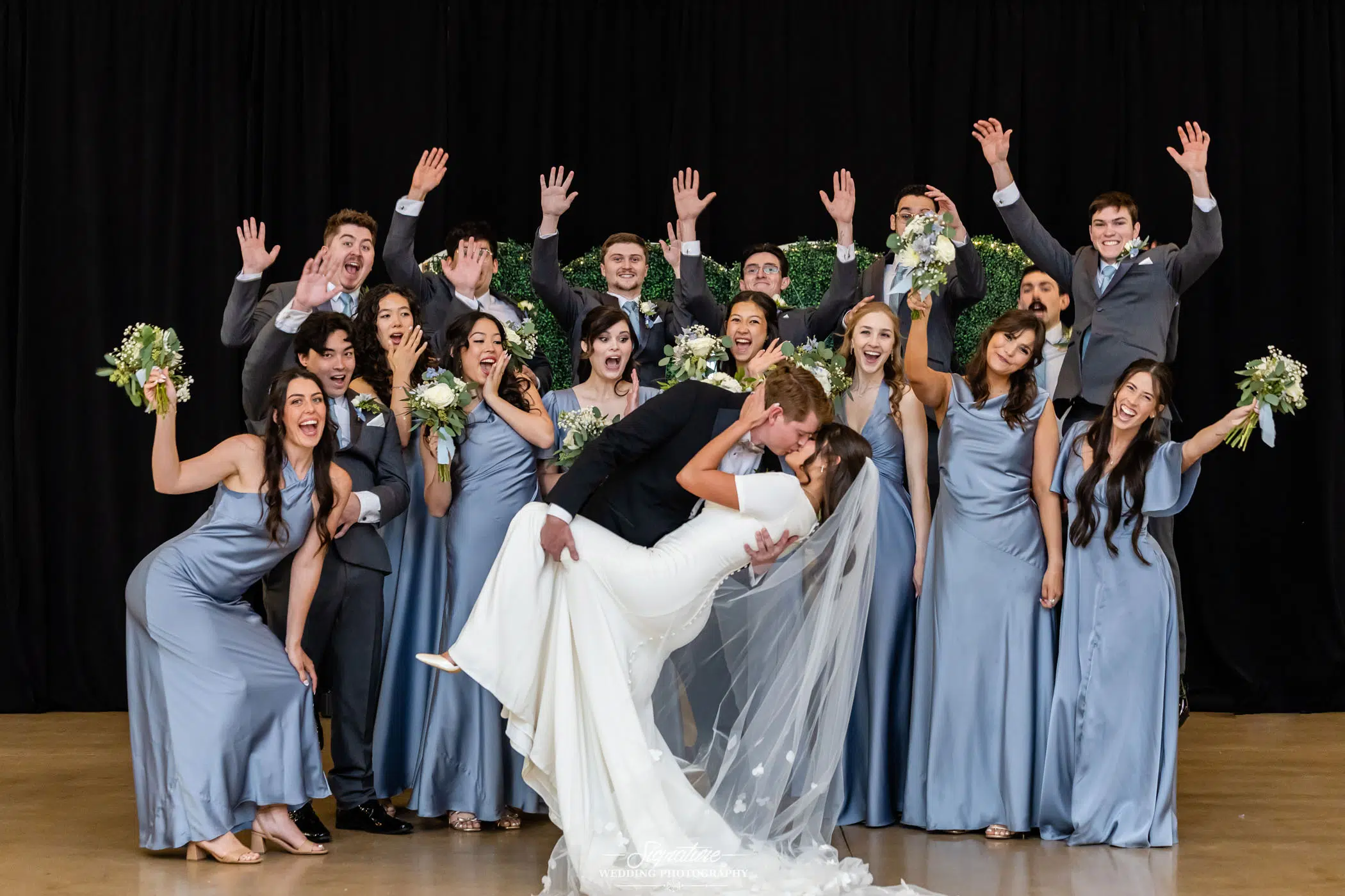Image by Signature Wedding Photography A bride and groom kiss in the center as bridesmaids in light blue dresses and groomsmen in gray suits cheer and pose enthusiastically against a black backdrop.
