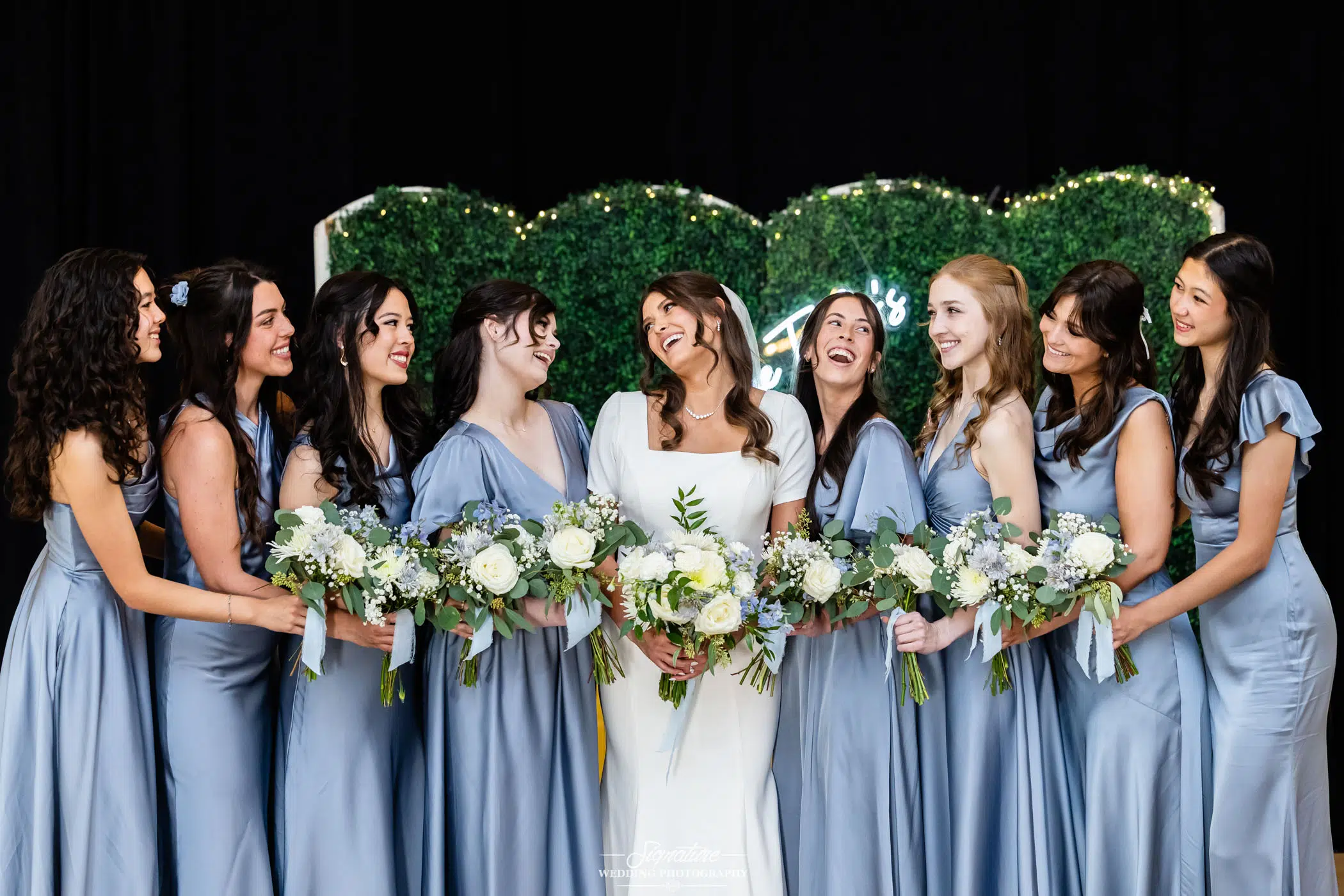 Image by Signature Wedding Photography A bride in a white dress stands smiling with seven bridesmaids in matching light blue dresses, all holding bouquets of white flowers, in front of a greenery backdrop with fairy lights.