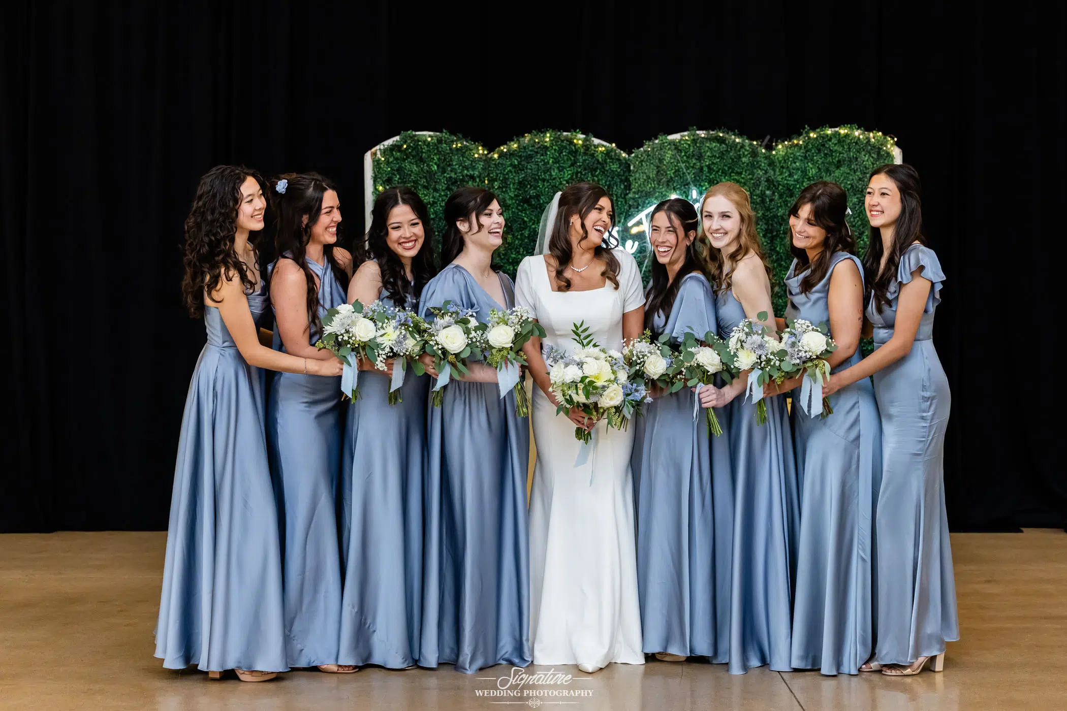 Image by Signature Wedding Photography A bride in a white dress stands with eight bridesmaids in matching light blue dresses, all smiling, holding bouquets, and standing in front of a green leafy backdrop.