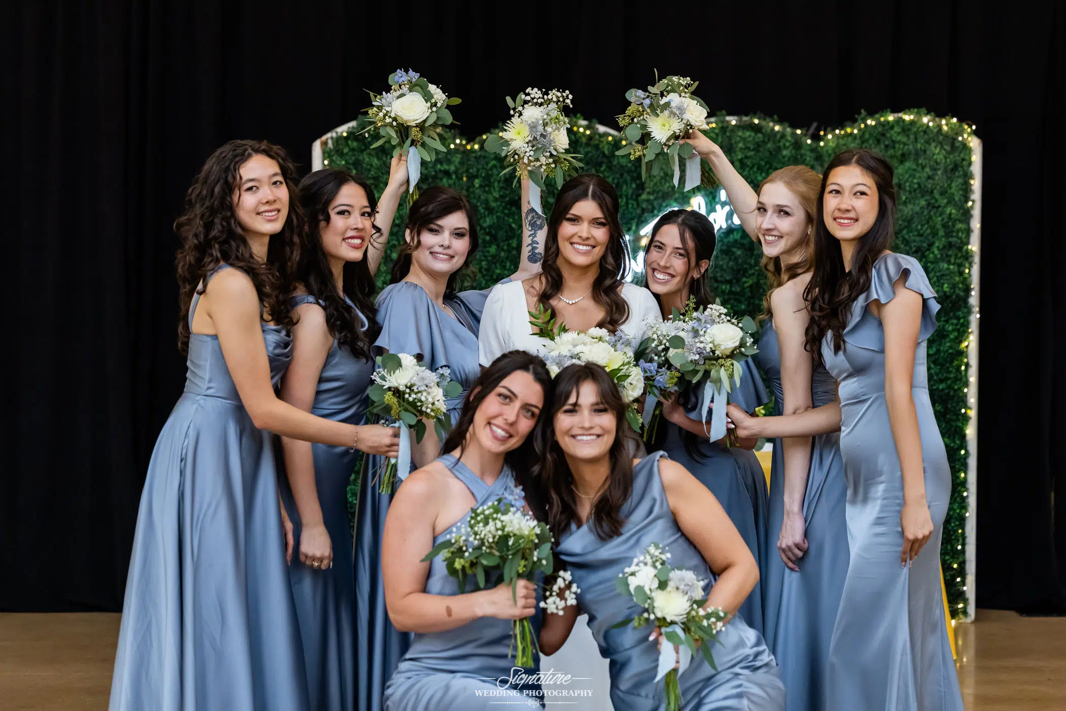 Image by Signature Wedding Photography A bride in white stands smiling, surrounded by eight bridesmaids in matching light blue dresses holding bouquets, posing together in front of greenery and fairy lights.