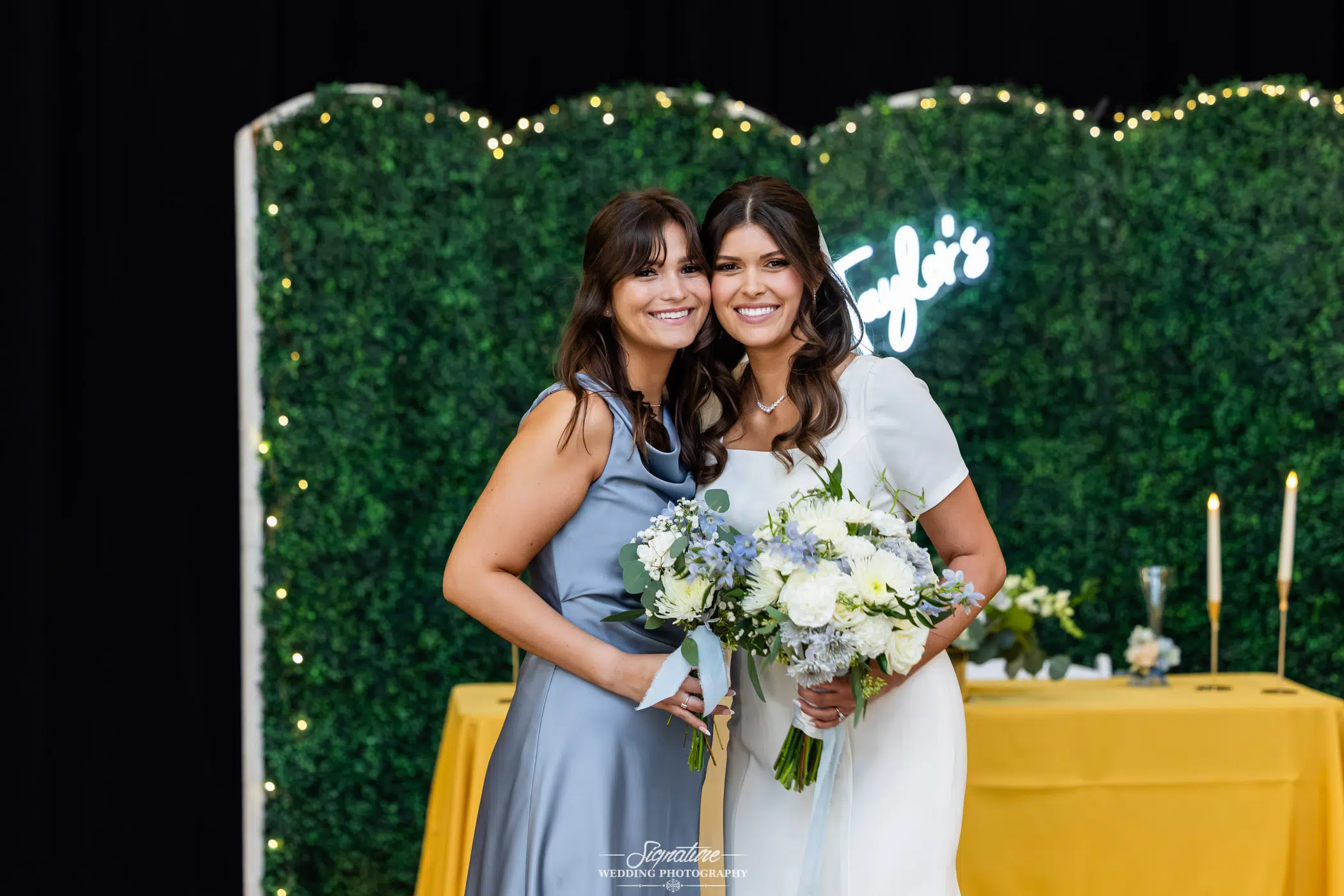 Image by Signature Wedding Photography Two women dressed formally, one in a light blue dress and the other in a white dress, smile and pose together holding bouquets in front of a green foliage backdrop with fairy lights and a yellow table.