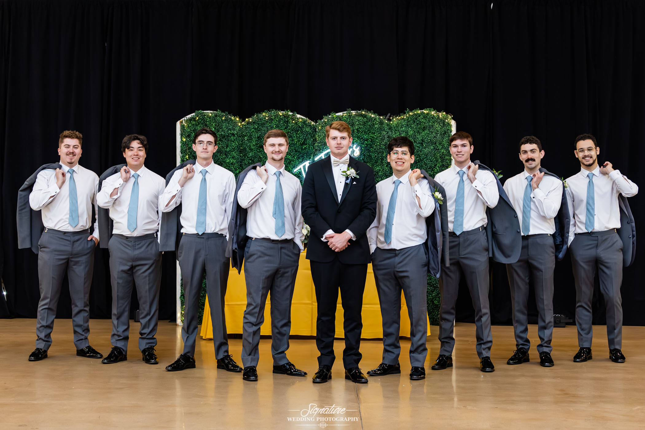 Image by Signature Wedding Photography A groom in a black suit stands in the center, surrounded by seven groomsmen in white shirts, light blue ties, and gray pants, holding suit jackets over their shoulders, posing in front of a green and yellow wedding backdrop.