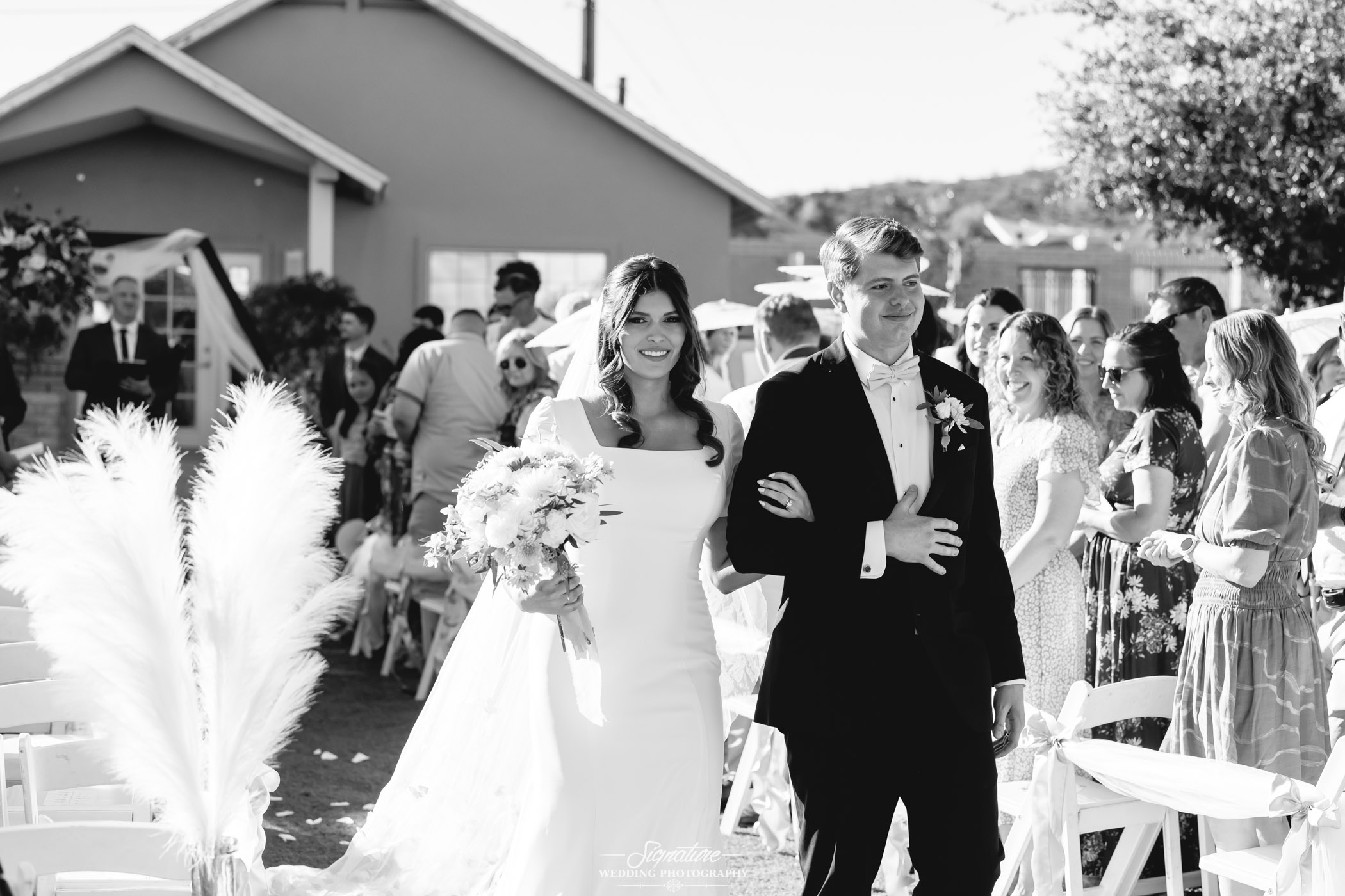 Image by Signature Wedding Photography A bride in a white dress holding a bouquet walks arm-in-arm with a man in a tuxedo down an outdoor aisle, surrounded by seated guests at a sunny wedding ceremony.