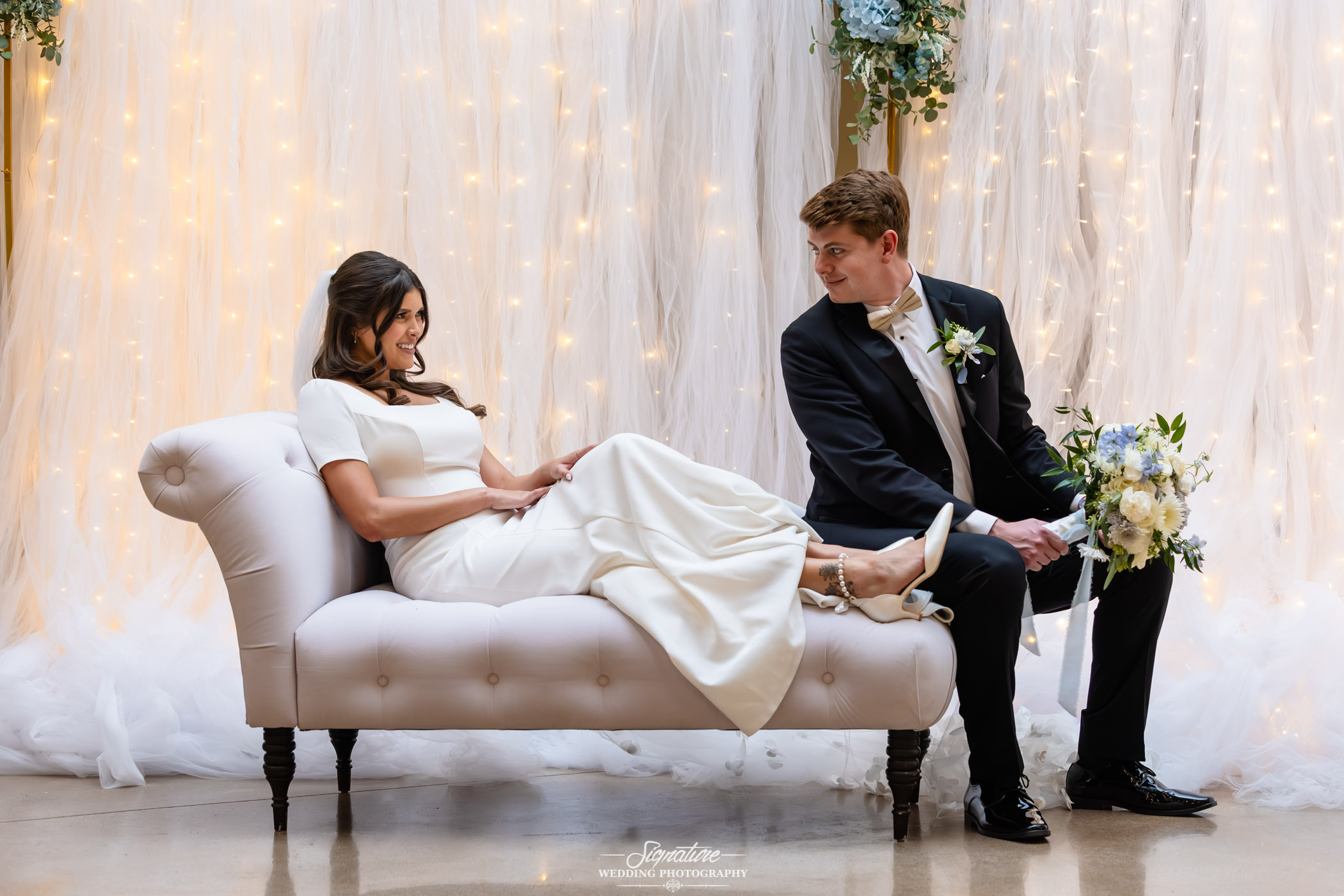 Image by Signature Wedding Photography A bride in a white dress reclines playfully on a chaise lounge, smiling at a groom in a black tuxedo who is placing a shoe on her foot, surrounded by soft white drapery and string lights with a floral arrangement nearby.