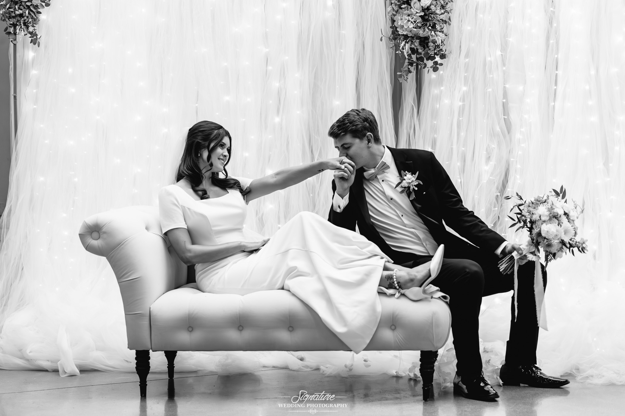 Image by Signature Wedding Photography A bride in a white dress sits on a sofa, smiling as the groom, in a suit, kneels and kisses her hand. Soft lights and draped fabric create a romantic, elegant atmosphere.