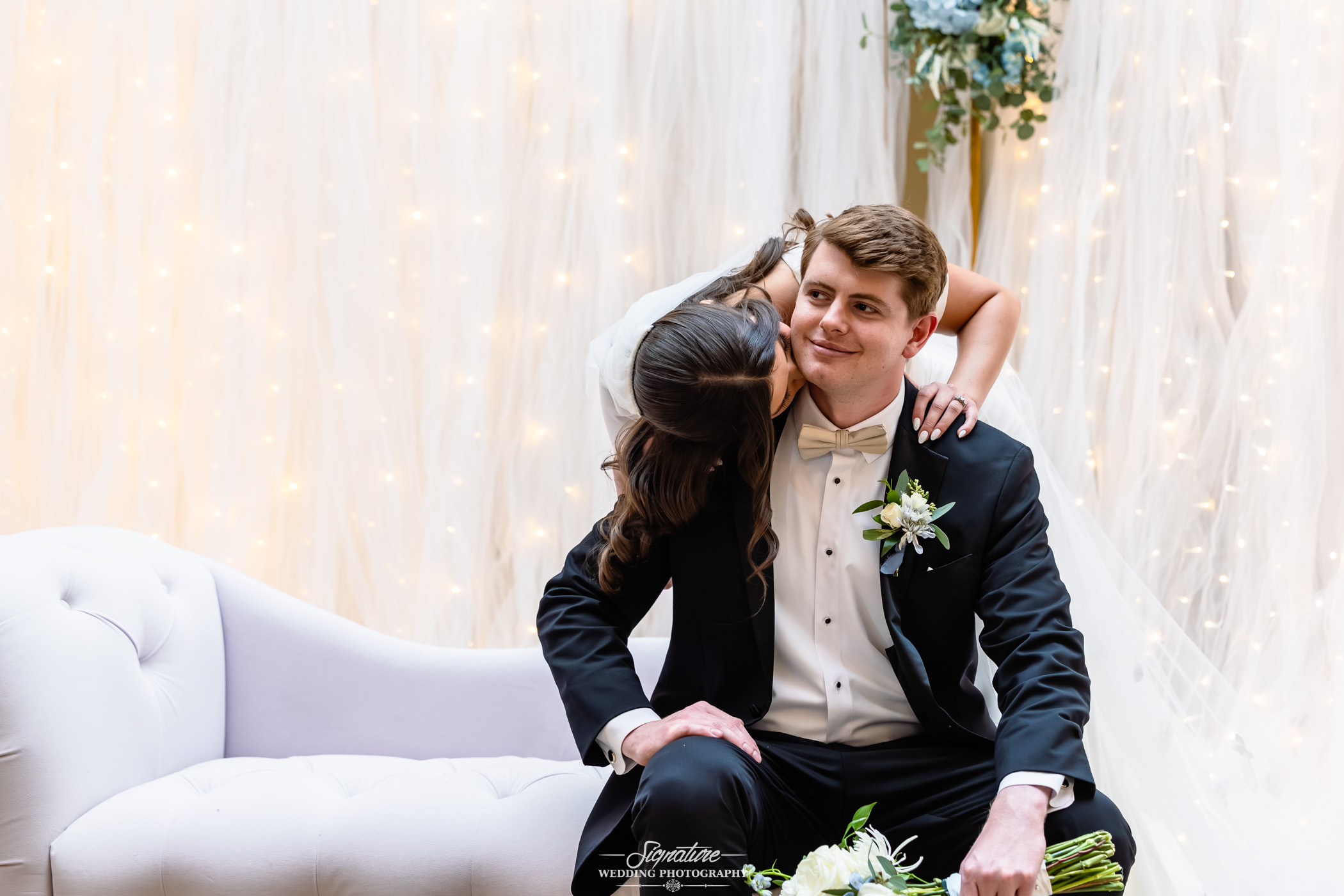 Image by Signature Wedding Photography A bride in a white dress leans over and kisses a seated groom on the cheek. The groom, in a black tuxedo with a white boutonniere, smiles. They are in front of a white draped backdrop with soft string lights.