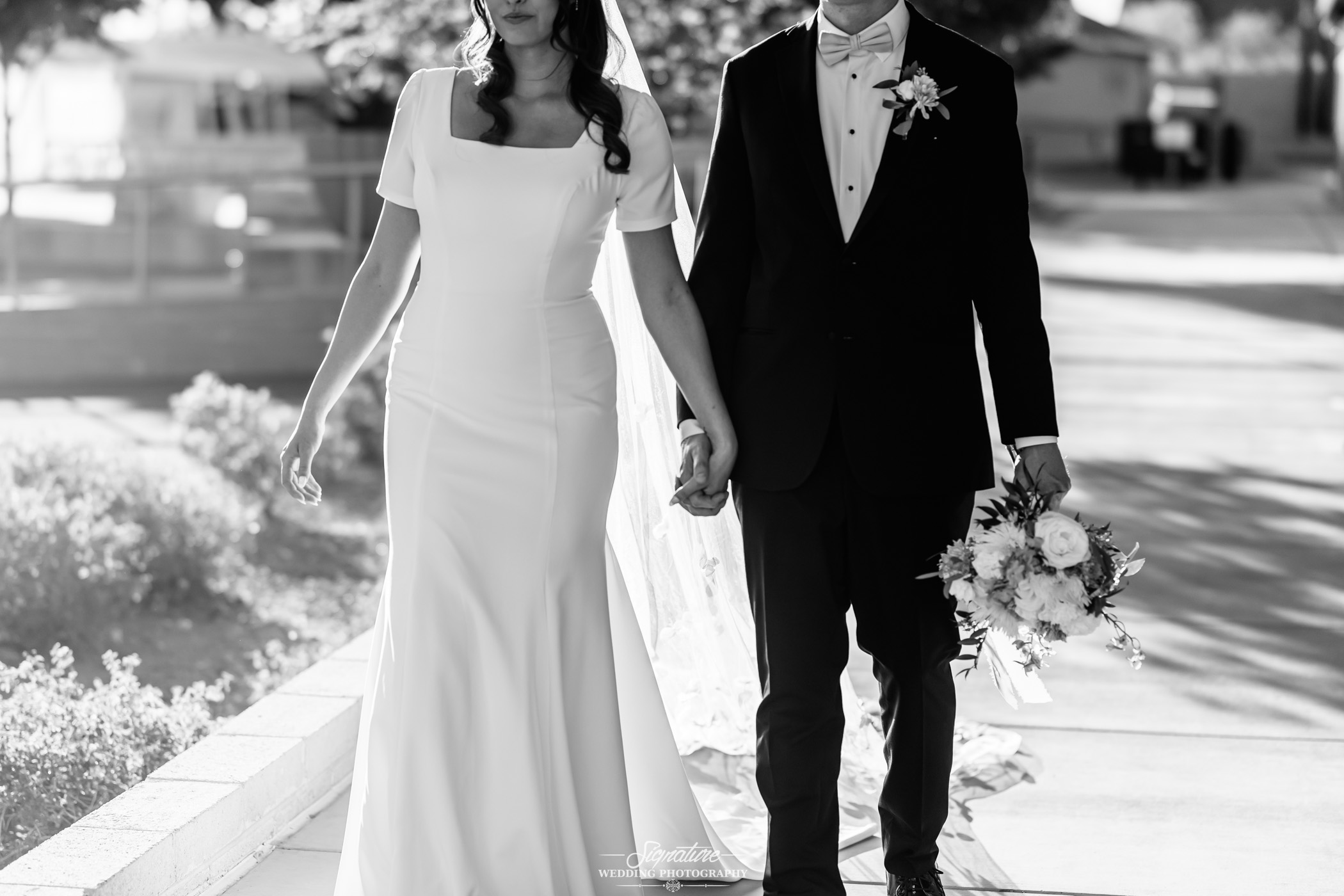 Image by Signature Wedding Photography A bride in a white dress and a groom in a suit walk hand in hand outdoors; the groom holds a bouquet of flowers. The image is in black and white.