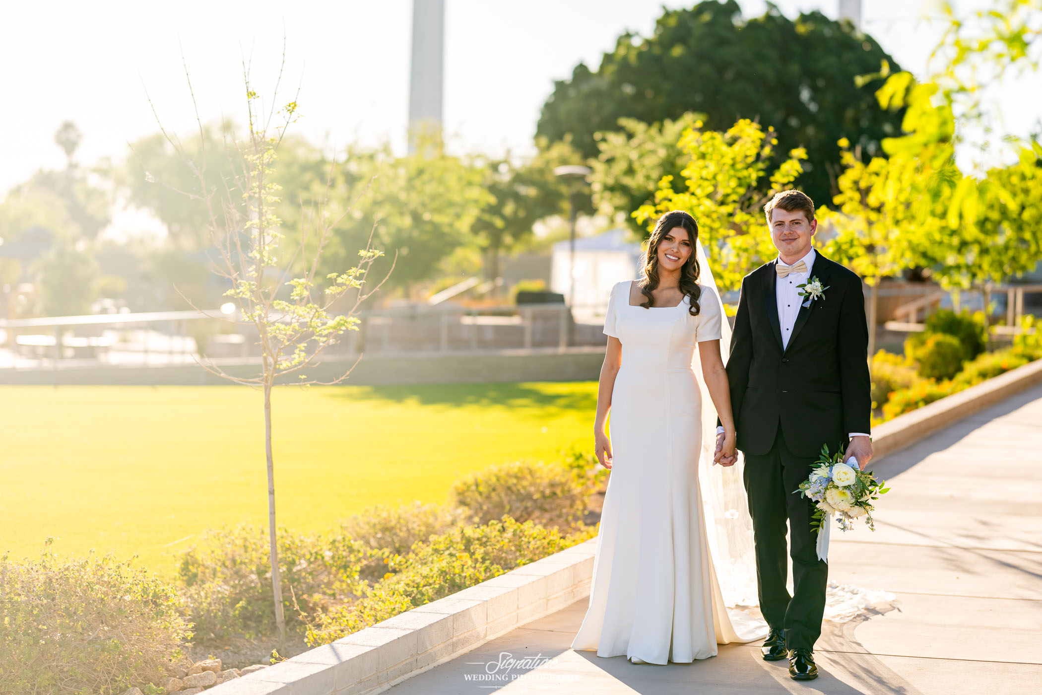 Image by Signature Wedding Photography A bride in a white dress and a groom in a black suit walk hand in hand outdoors on a sunny day, surrounded by green trees and grass, both smiling at the camera. The groom holds a bouquet.