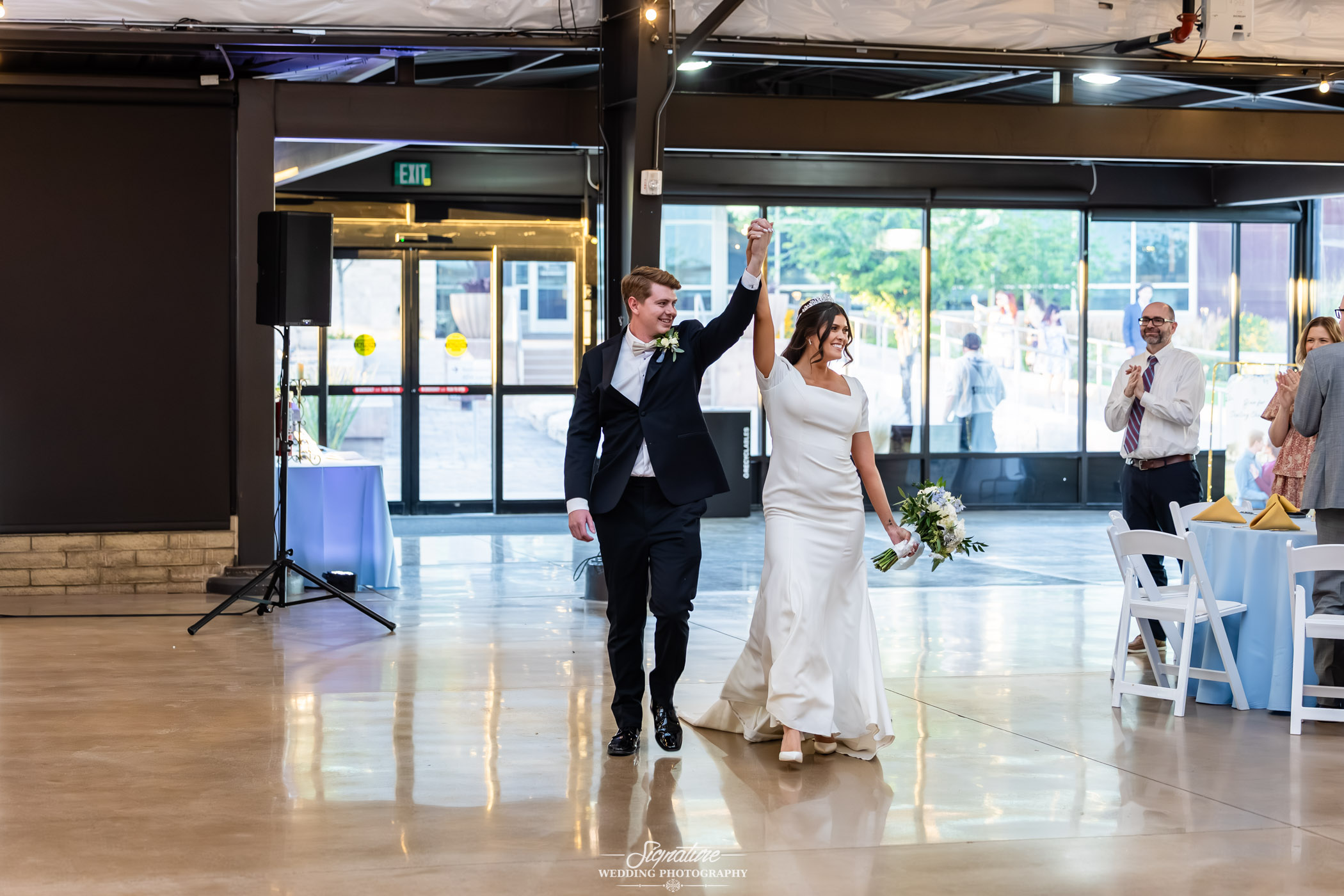 Image by Signature Wedding Photography A newlywed couple walks into a bright reception hall, holding hands with raised arms and smiling. The bride wears a white dress, and the groom is in a black suit. Guests applaud in the background.