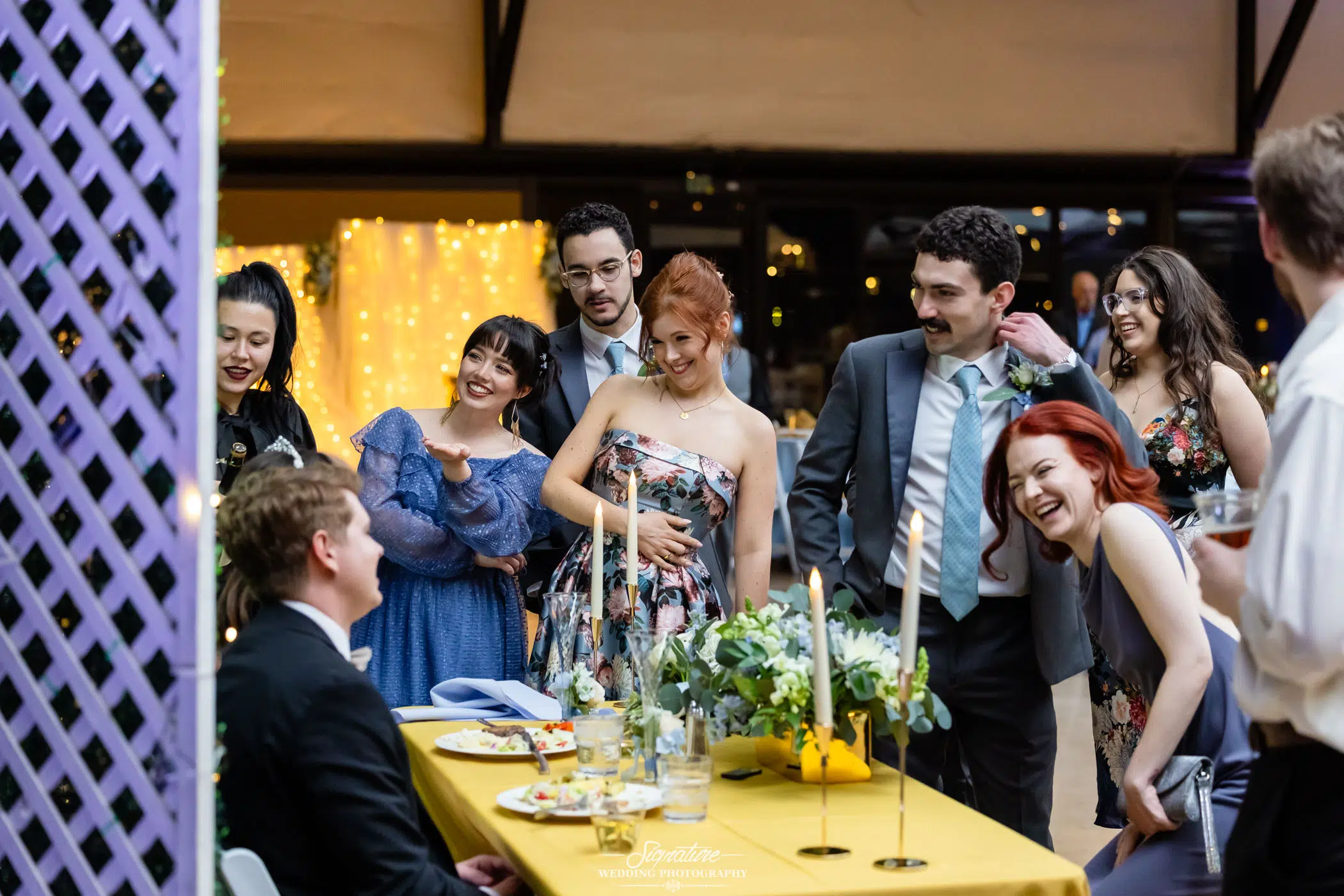 Image by Signature Wedding Photography A group of well-dressed people stand and laugh together around a table decorated with candles and flowers at a lively indoor celebration, while a man in a tuxedo sits smiling at the group.