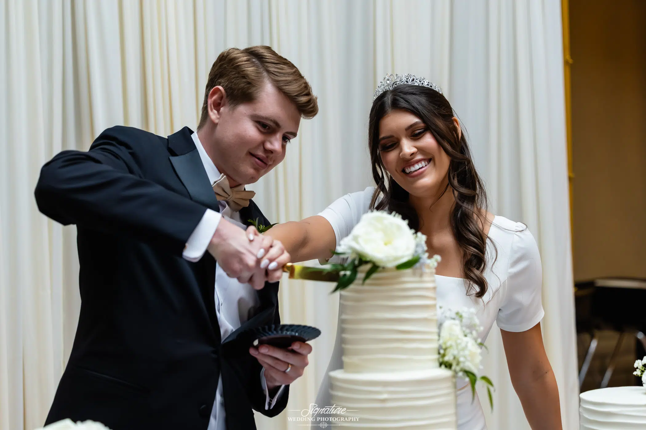 Image by Signature Wedding Photography A bride and groom, both smiling, cut a white tiered wedding cake together. The bride wears a white dress and tiara, and the groom is in a black tuxedo with a bow tie. White flowers decorate the cake.