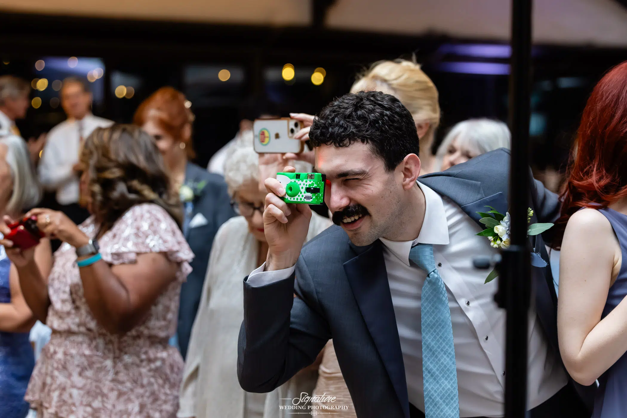 Image by Signature Wedding Photography A man in a suit and tie holds a green disposable camera up to his eye, smiling at an event. Other guests, some with cameras and phones, stand in the background, dressed in formal attire.