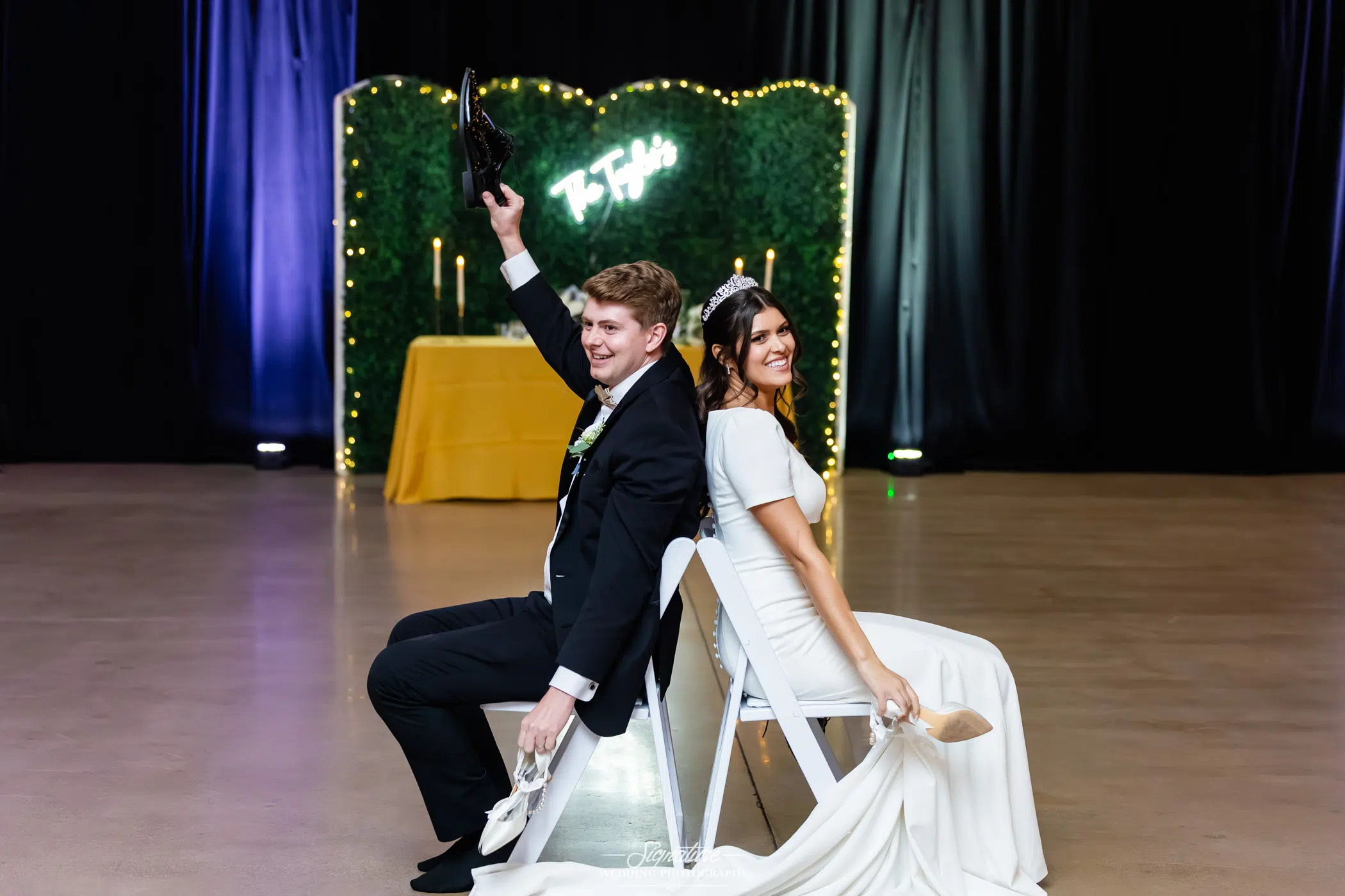 Image by Signature Wedding Photography A bride and groom sit back-to-back on white chairs at their wedding, each holding up a shoe in the "shoe game." The groom wears a black suit; the bride is in a white dress. A green backdrop with lights and their name is behind them.