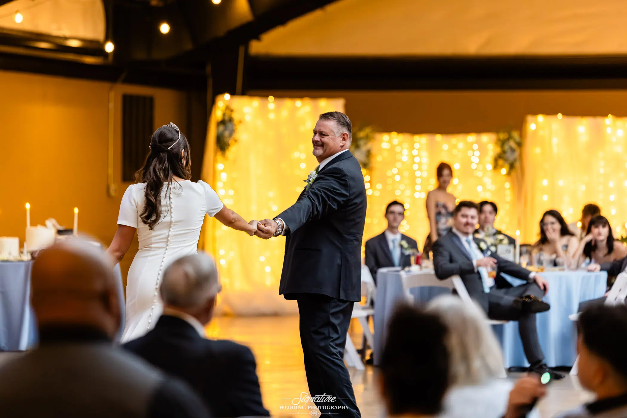 Image by Signature Wedding Photography A bride and groom dance together in a warmly lit reception hall, smiling, with guests seated at decorated tables watching them. Twinkling string lights and yellow drapery create a festive atmosphere in the background.