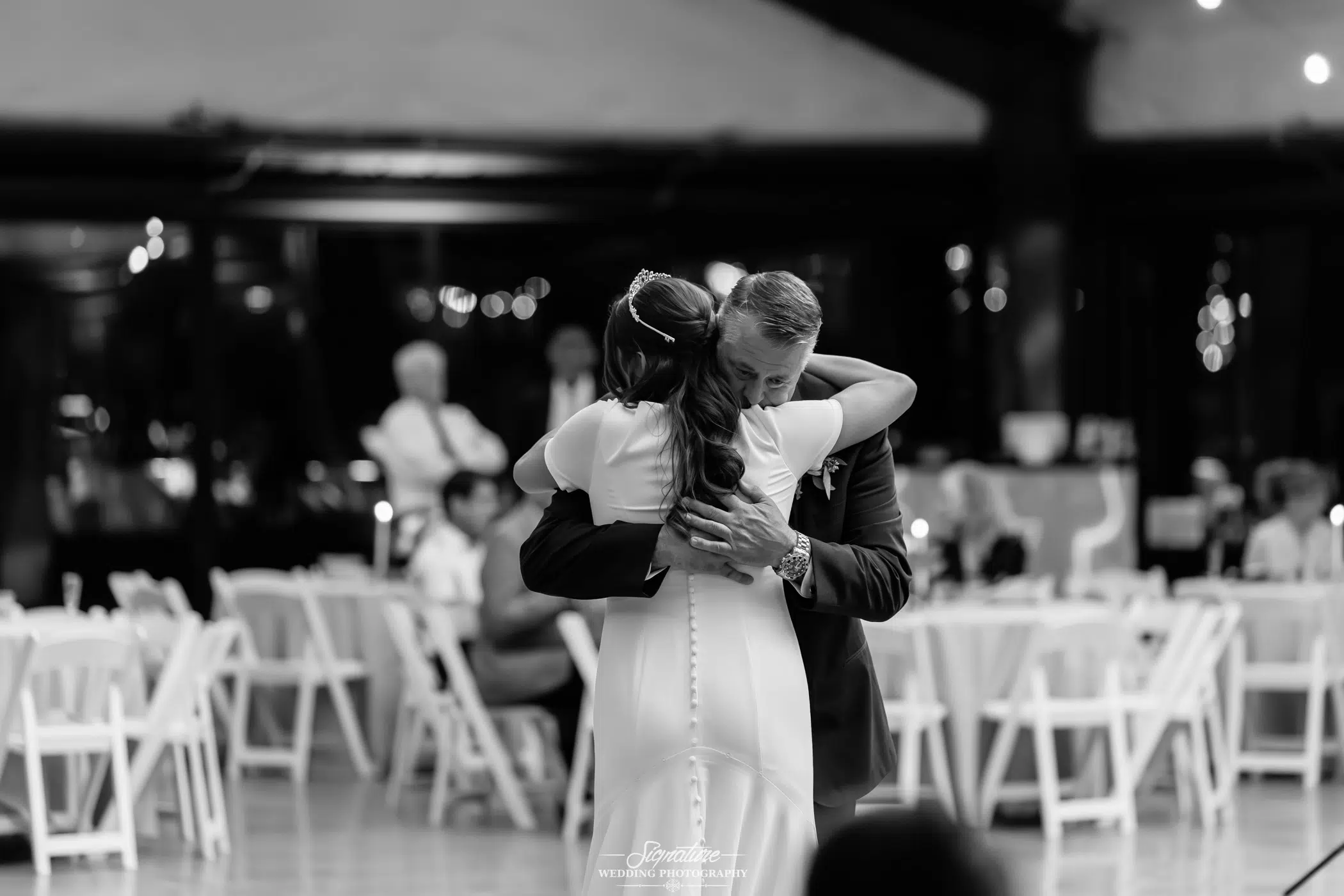 Image by Signature Wedding Photography A bride in a white dress hugs an older man, likely her father, during a dance at a wedding reception in a room with white chairs and tables. The background is softly blurred. The image is in black and white.