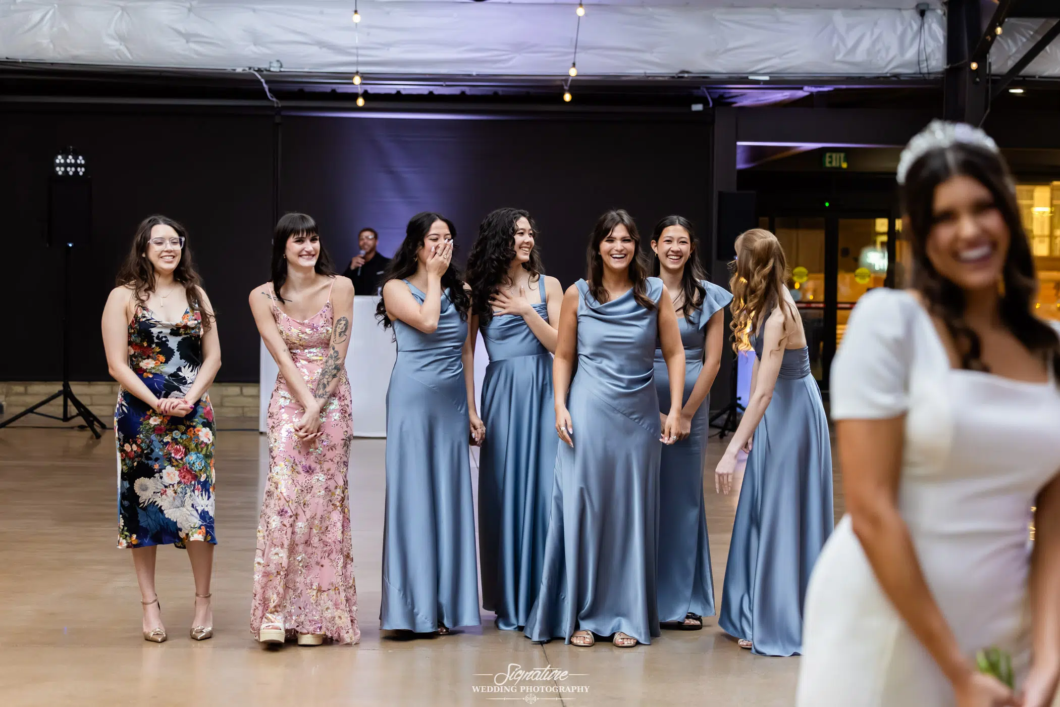 Image by Signature Wedding Photography A group of women, some in blue satin dresses and others in floral dresses, stand smiling and laughing indoors as a bride in a white gown prepares to throw her bouquet in the foreground.