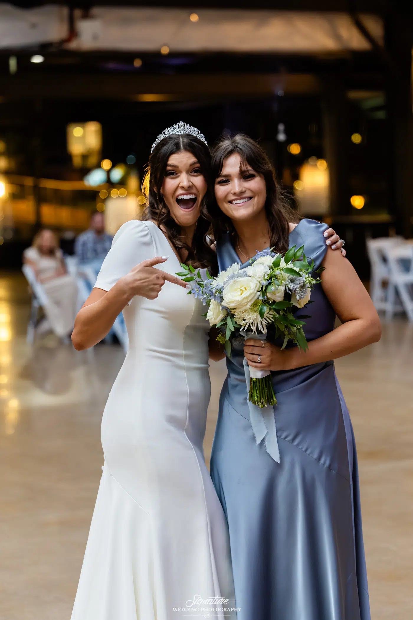 Image by Signature Wedding Photography Two women stand together, smiling; one in a white dress and tiara excitedly points at the other, who wears a light blue gown and holds a bouquet. They appear happy at a wedding reception.