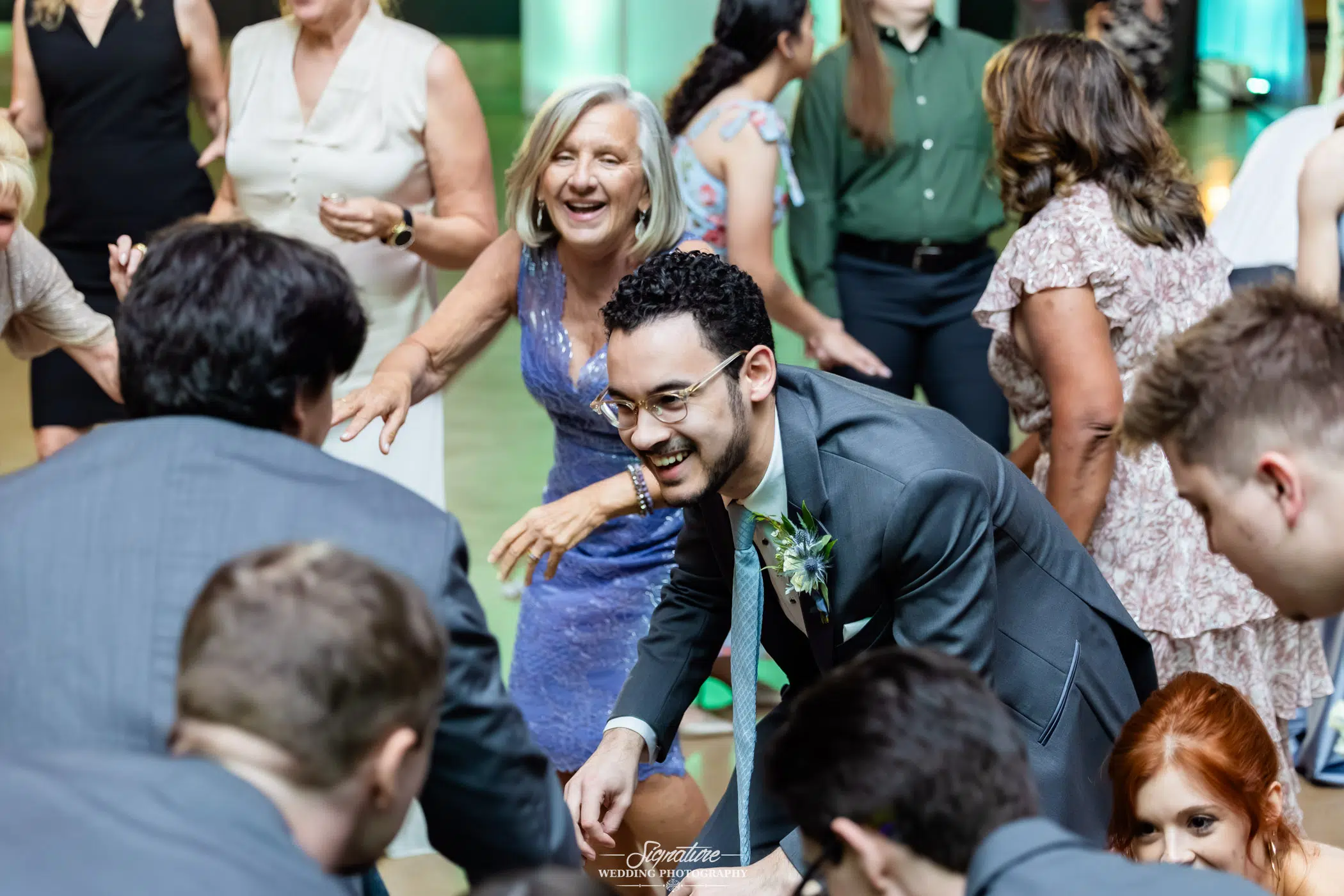 Image by Signature Wedding Photography A smiling man in a gray suit and glasses bends forward while dancing, surrounded by other laughing guests on a crowded dance floor at a celebration. A woman in a blue dress smiles behind him.