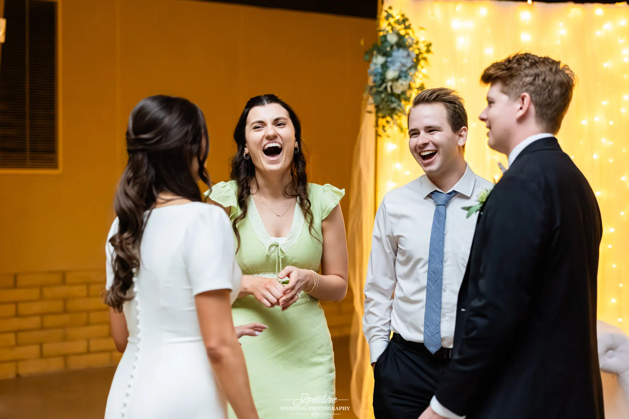 Image by Signature Wedding Photography Four young adults, two women and two men, stand together indoors, laughing and talking. The background features warm yellow lighting and a decorative floral arrangement. The atmosphere appears festive and joyful.