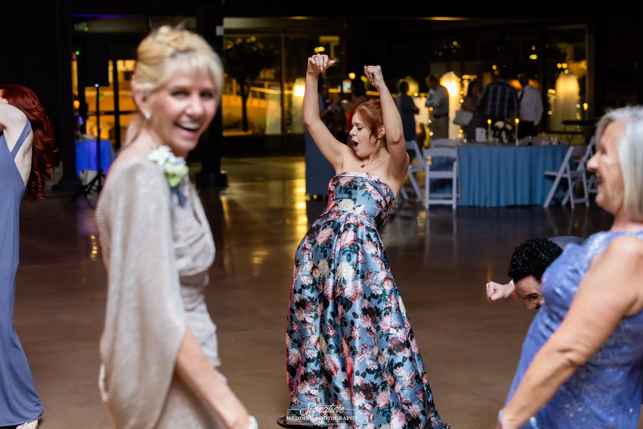 Image by Signature Wedding Photography A woman in a floral dress dances energetically with her arms raised at an indoor event, while other people nearby smile and enjoy themselves. The atmosphere is lively and festive.