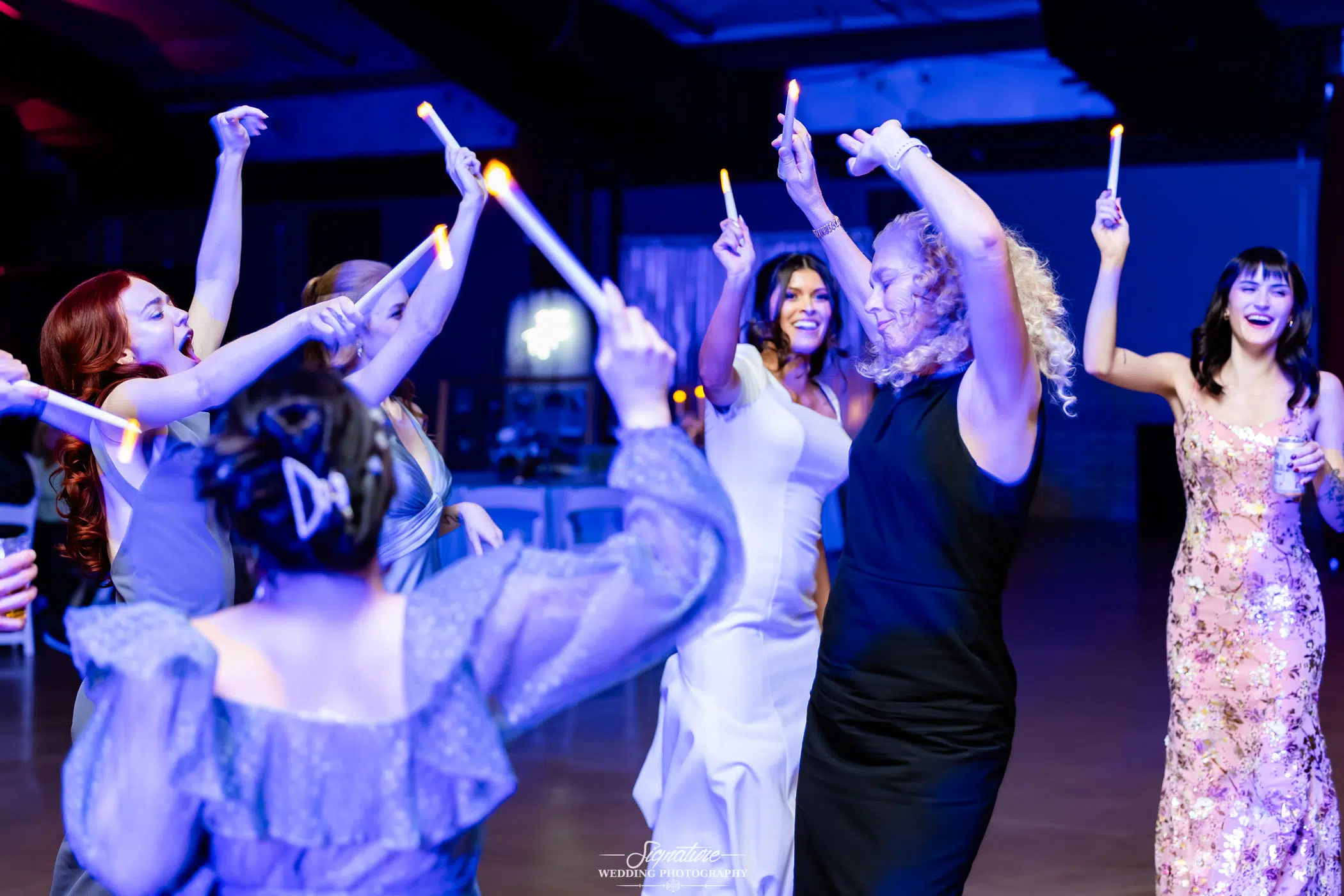 Image by Signature Wedding Photography A group of women in formal dresses dance energetically with glow sticks at a lively indoor event, smiling and celebrating together under colorful lighting.