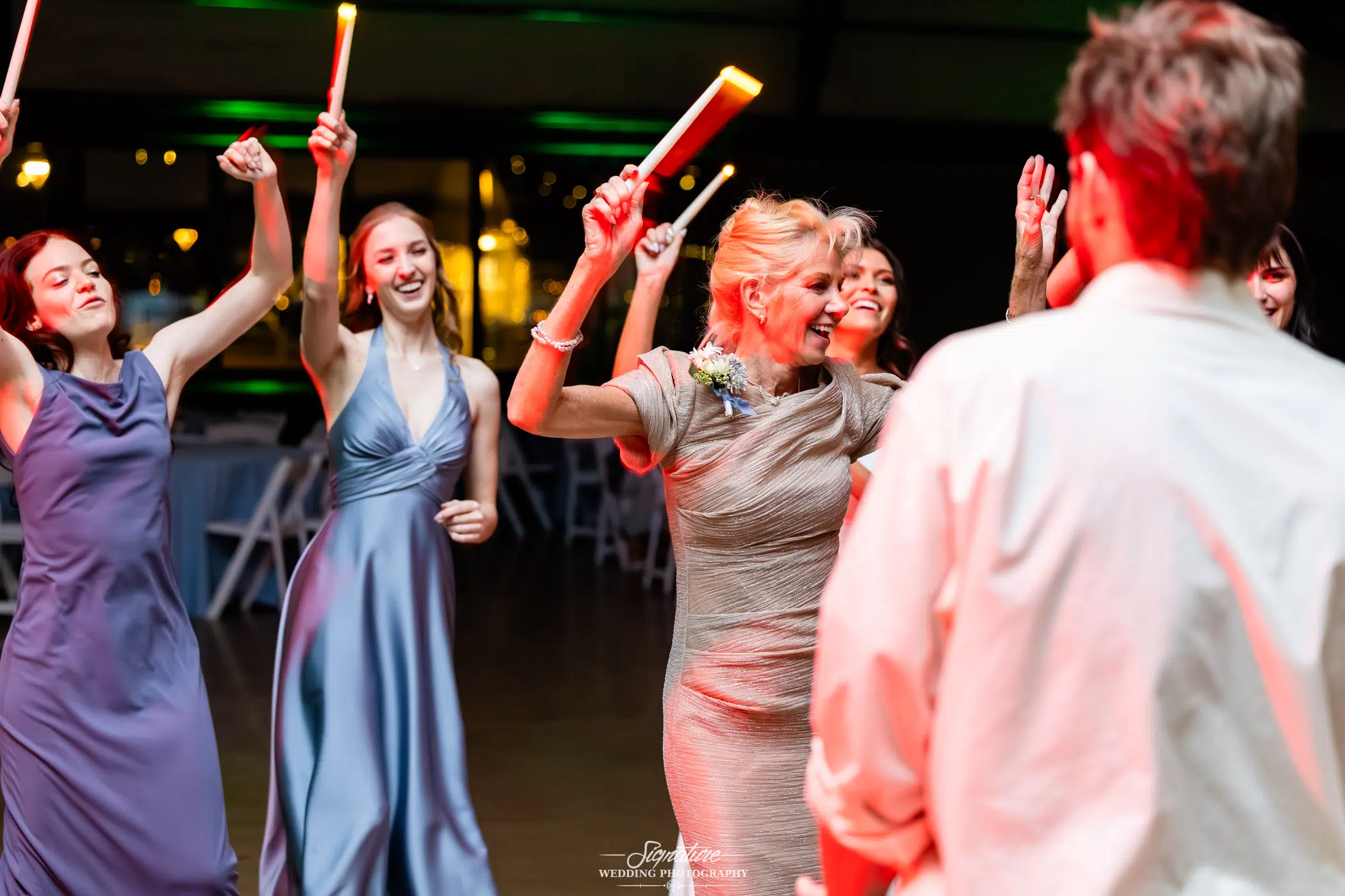 Image by Signature Wedding Photography A group of women in formal dresses dance joyfully with glow sticks at an indoor event, while a man in a white shirt faces them from the foreground.
