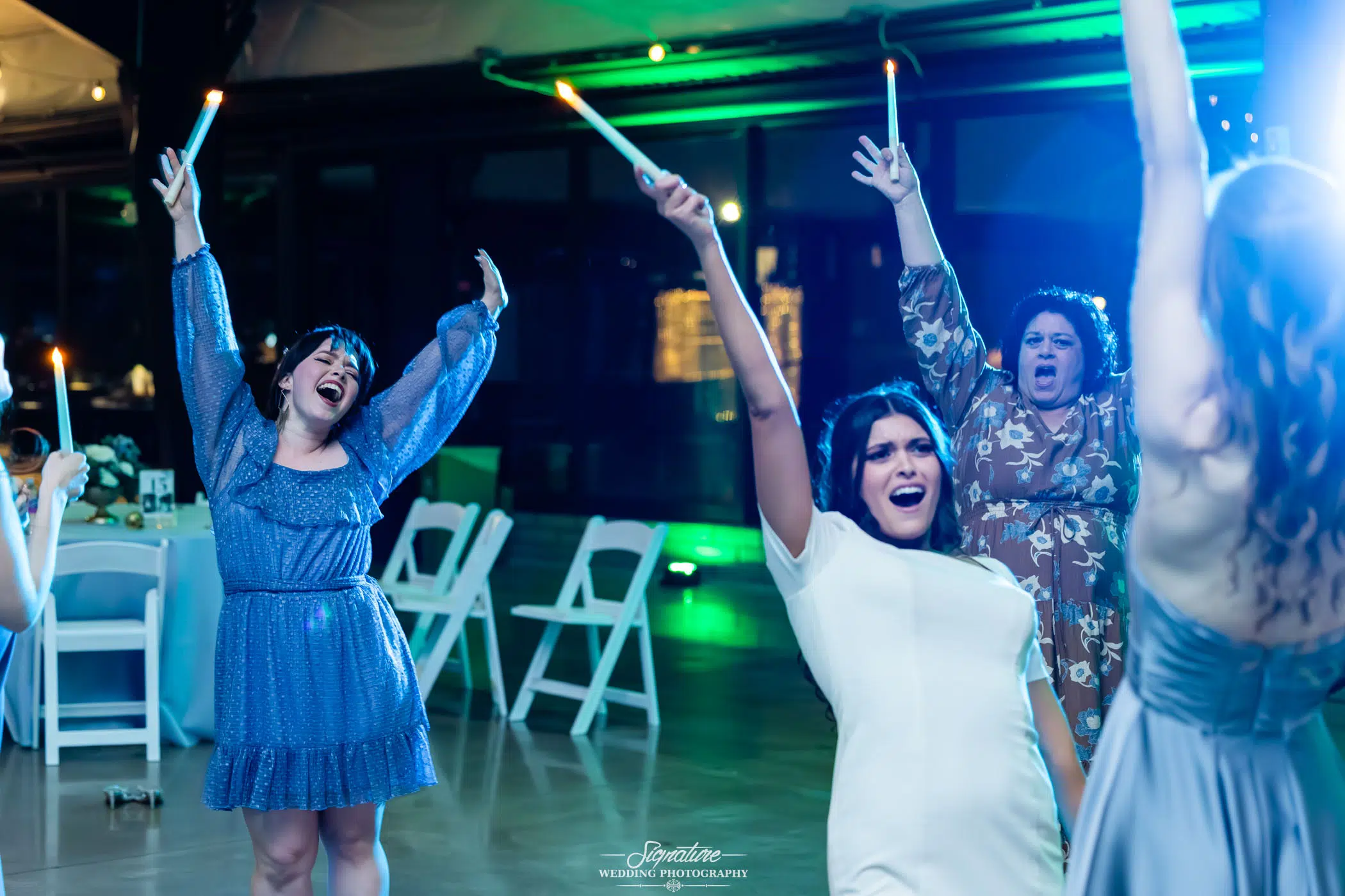 Image by Signature Wedding Photography A group of women joyfully dance indoors, holding glowing sticks in the air. They are smiling, with their arms raised, surrounded by empty chairs and green lighting, creating a lively and festive atmosphere.