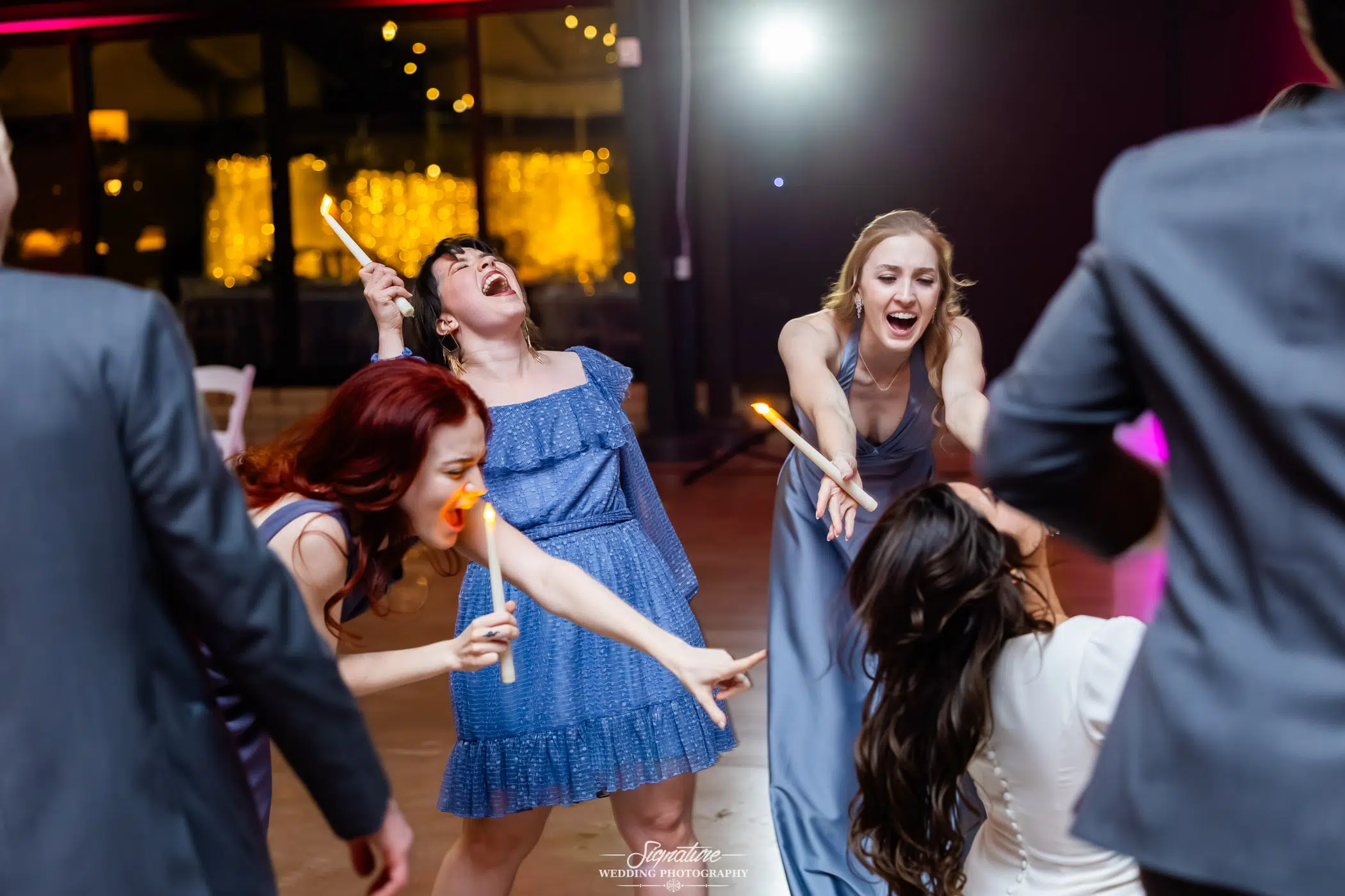 Image by Signature Wedding Photography Four women in colorful dresses laugh and sing while holding glow sticks on a dance floor at a lively indoor celebration with warm lights in the background.