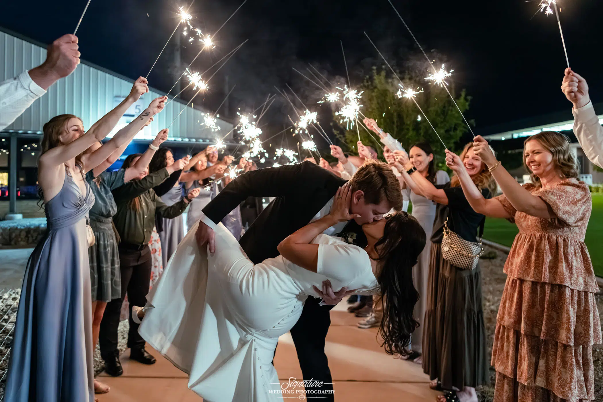 Image by Signature Wedding Photography A bride and groom kiss as the groom dips the bride. They are surrounded by guests holding sparklers in celebration at night, creating a joyful and festive atmosphere.