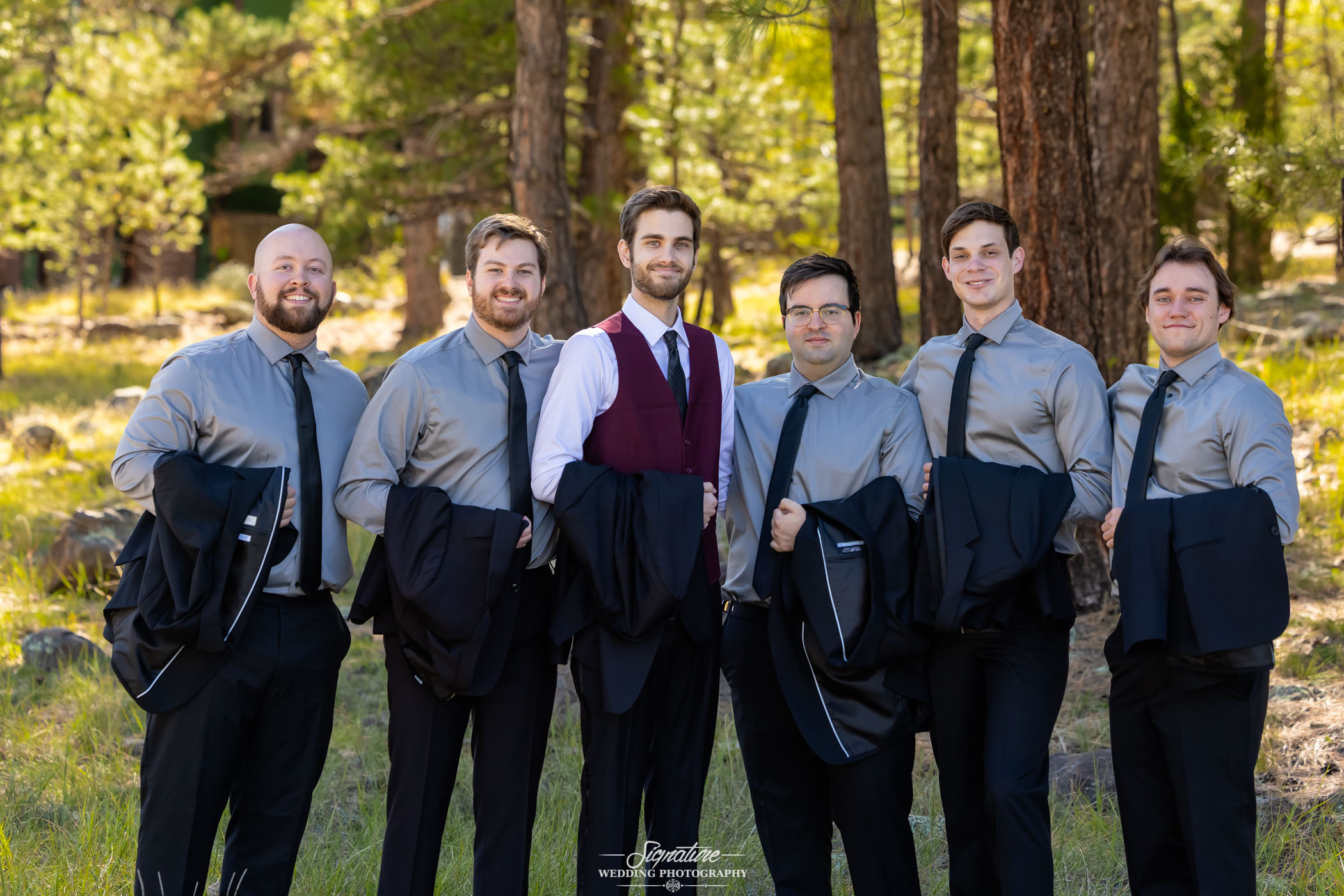 Photo by Tavits Photography Six men stand outdoors in a forested area, five wearing gray shirts and black ties, holding suit jackets, and one in a burgundy vest with a white shirt. All are smiling and posing for a group photo.