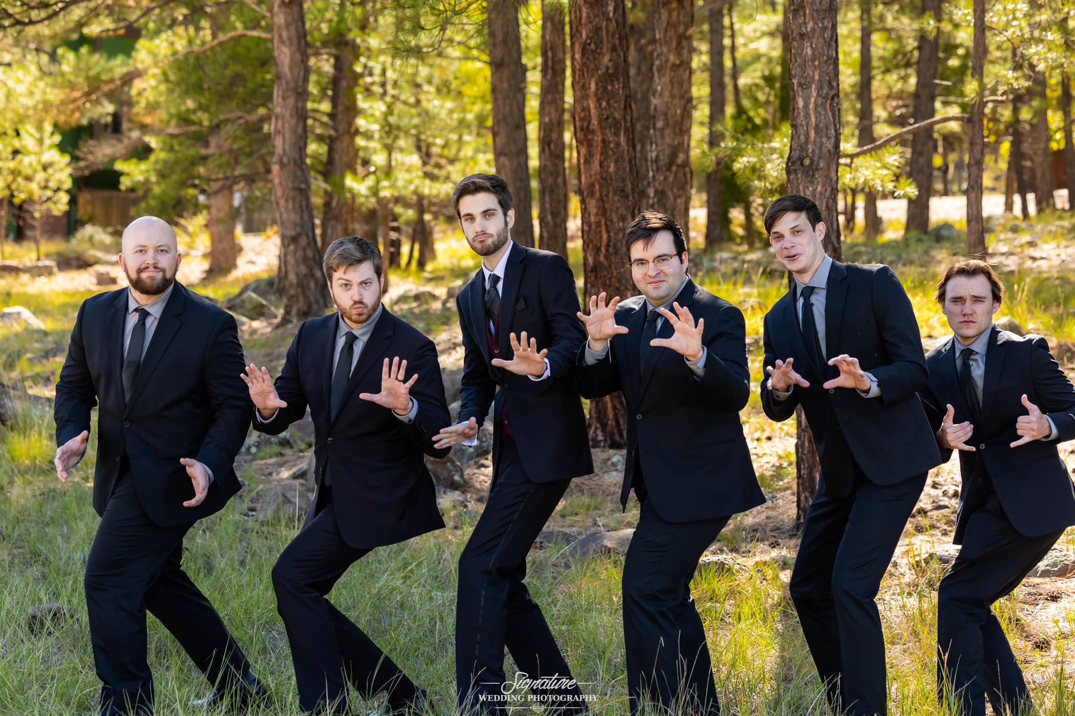 Photo by Tavits Photography Six men in black suits and ties stand outdoors in a pine forest, striking playful, dramatic poses with their hands raised as if in mid-action, while smiling and looking at the camera.