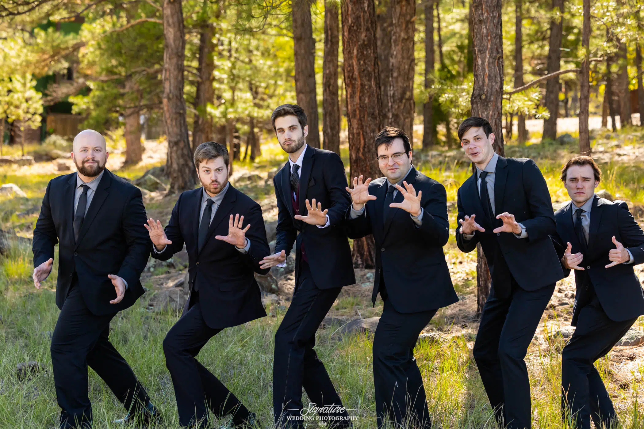 Photo by Tavits Photography Six men in black suits and ties stand outdoors in a pine forest, striking playful, dramatic poses with their hands raised as if in mid-action, while smiling and looking at the camera.