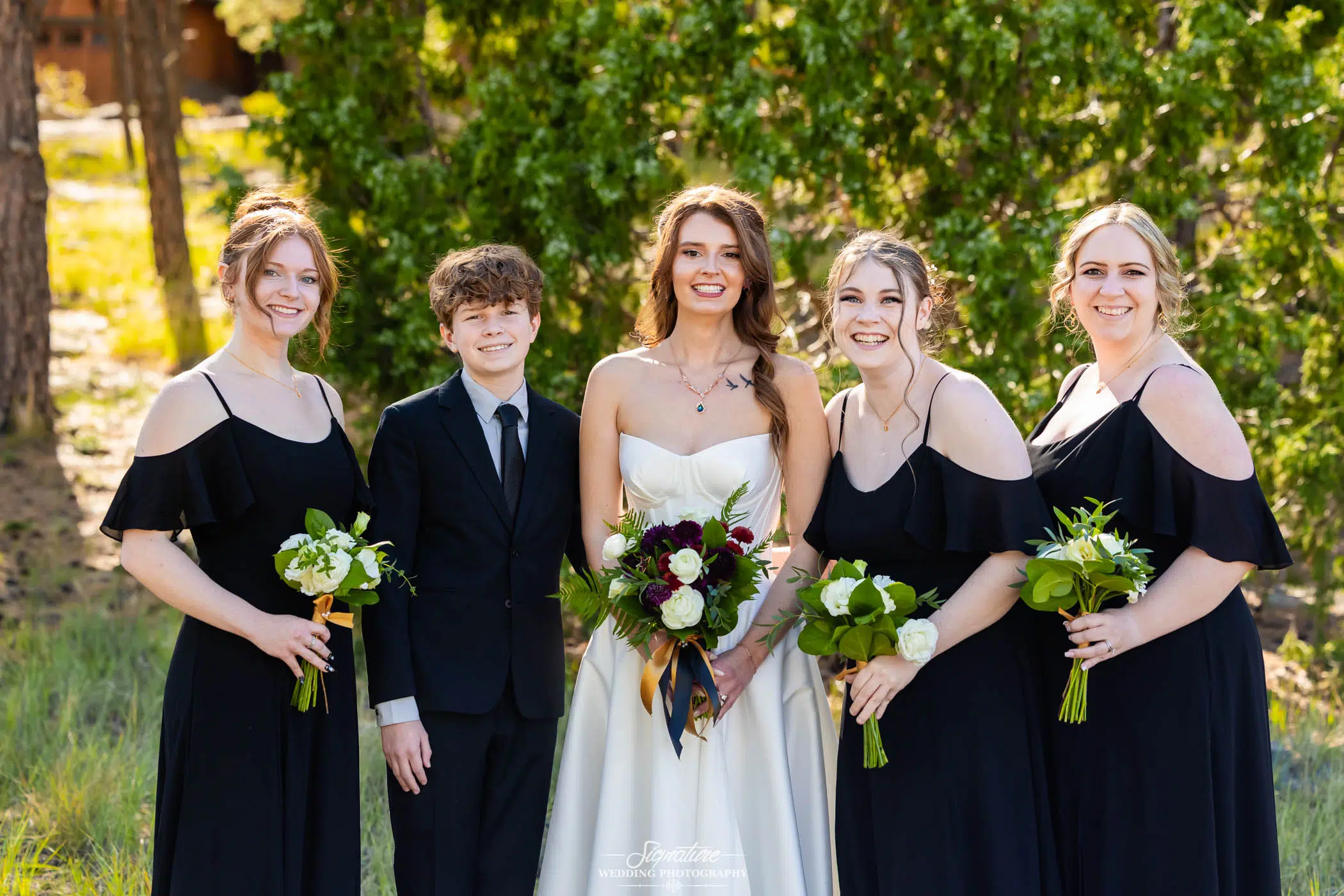 Photo by Tavits Photography Five people stand outdoors, dressed formally. The person in the center wears a white dress and holds a bouquet of flowers; the others wear black outfits and also hold bouquets. Green trees and grass are visible in the background.
