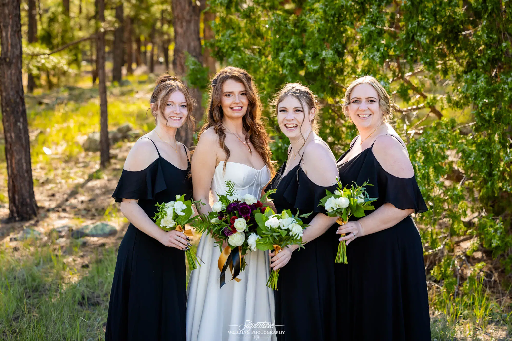 Photo by Tavits Photography Four women in black dresses, holding bouquets, stand outdoors in a sunlit forest. One woman in a white dress, likely the bride, stands in the center, all smiling at the camera.