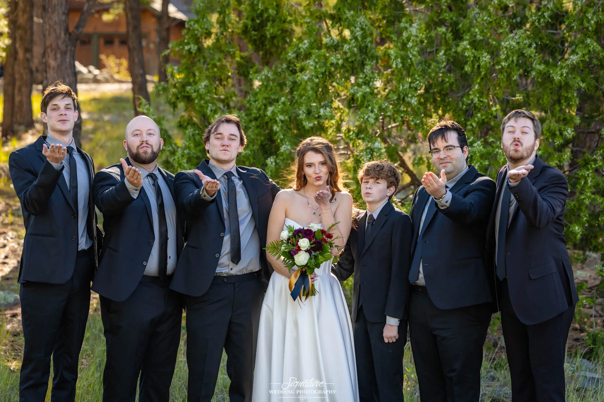Photo by Tavits Photography A bride in a white dress holding a bouquet stands outside with six men in suits, all blowing kisses toward the camera, surrounded by green trees and sunlight.