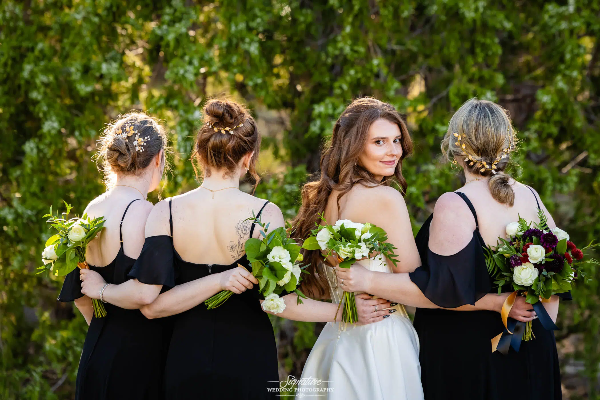 Photo by Tavits Photography Four women in formal dresses stand with their backs to the camera, arms linked. One woman, in a white dress, turns to face the camera, smiling. Each holds a bouquet; trees fill the background.