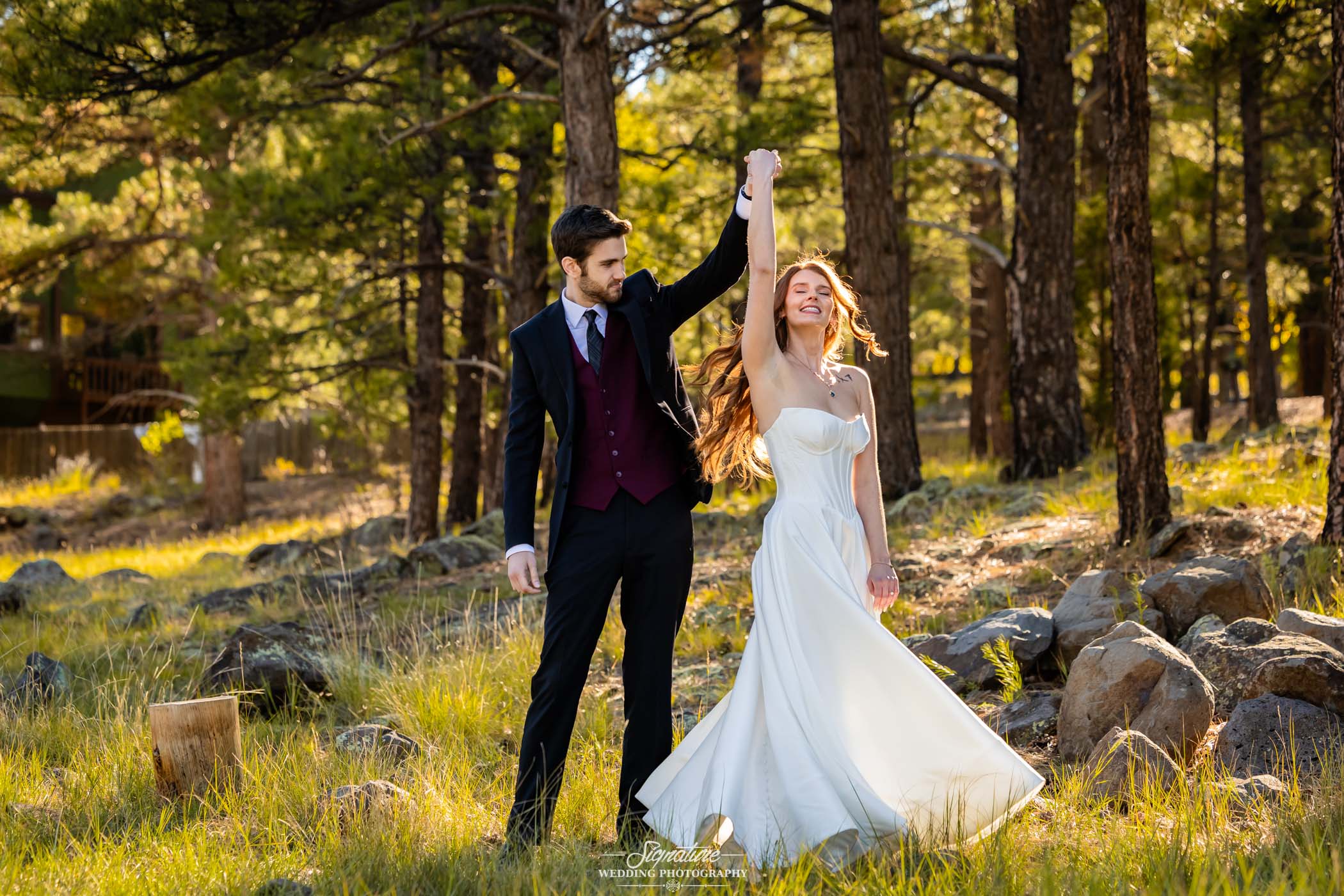 Photo by Tavits Photography A couple dances outdoors in a sunlit forest. The man, in a dark suit and burgundy vest, lifts the woman's hand as she twirls in a white wedding dress, smiling amid tall trees and grass-covered ground.